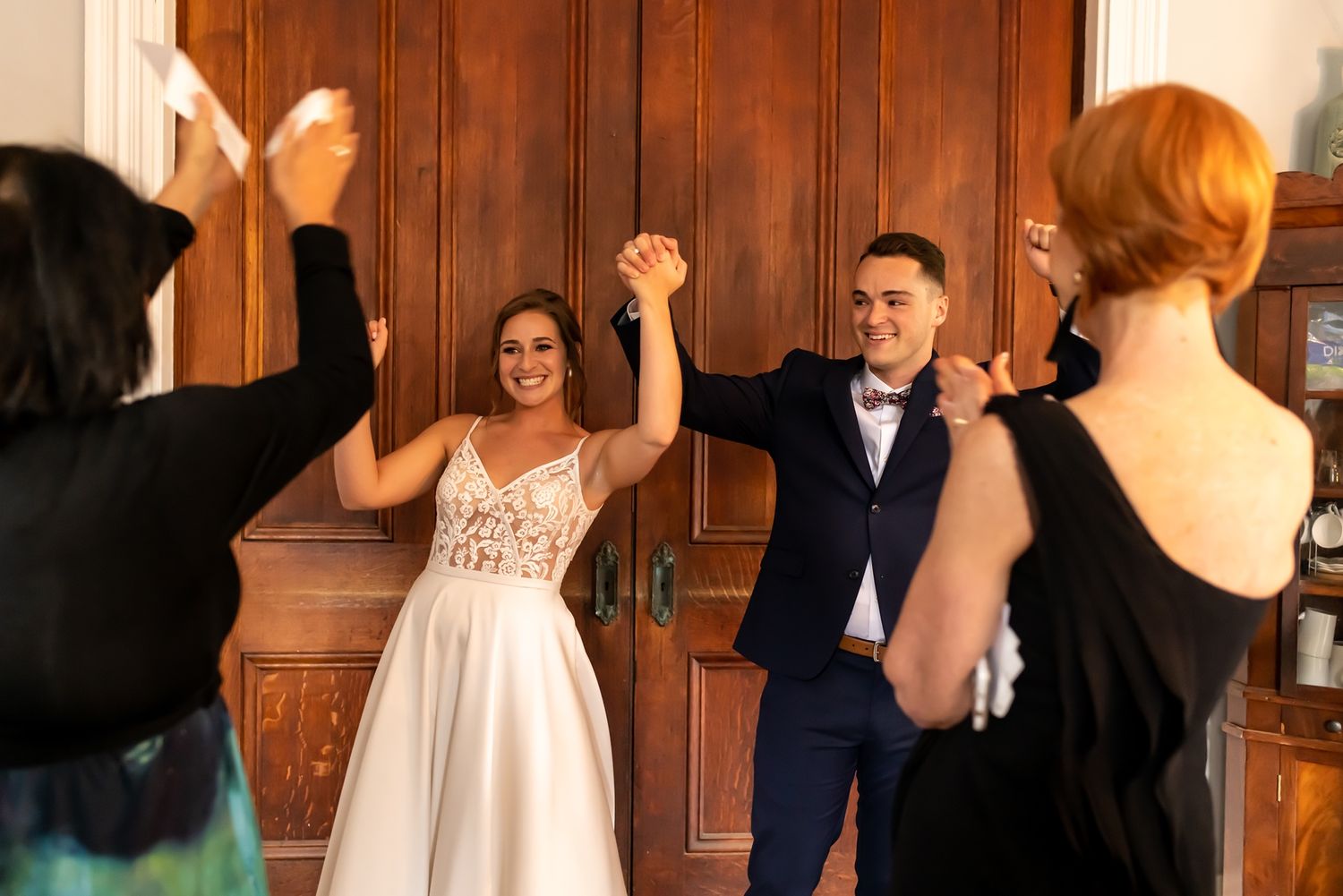Wedding couple holding hands and raising their arms in celebration while surrounded by guests in dark attire.