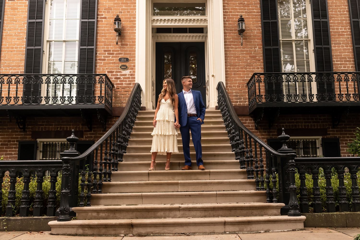 A couple stands on the stone steps of a historic brick townhouse with black iron railings and traditional architecture.