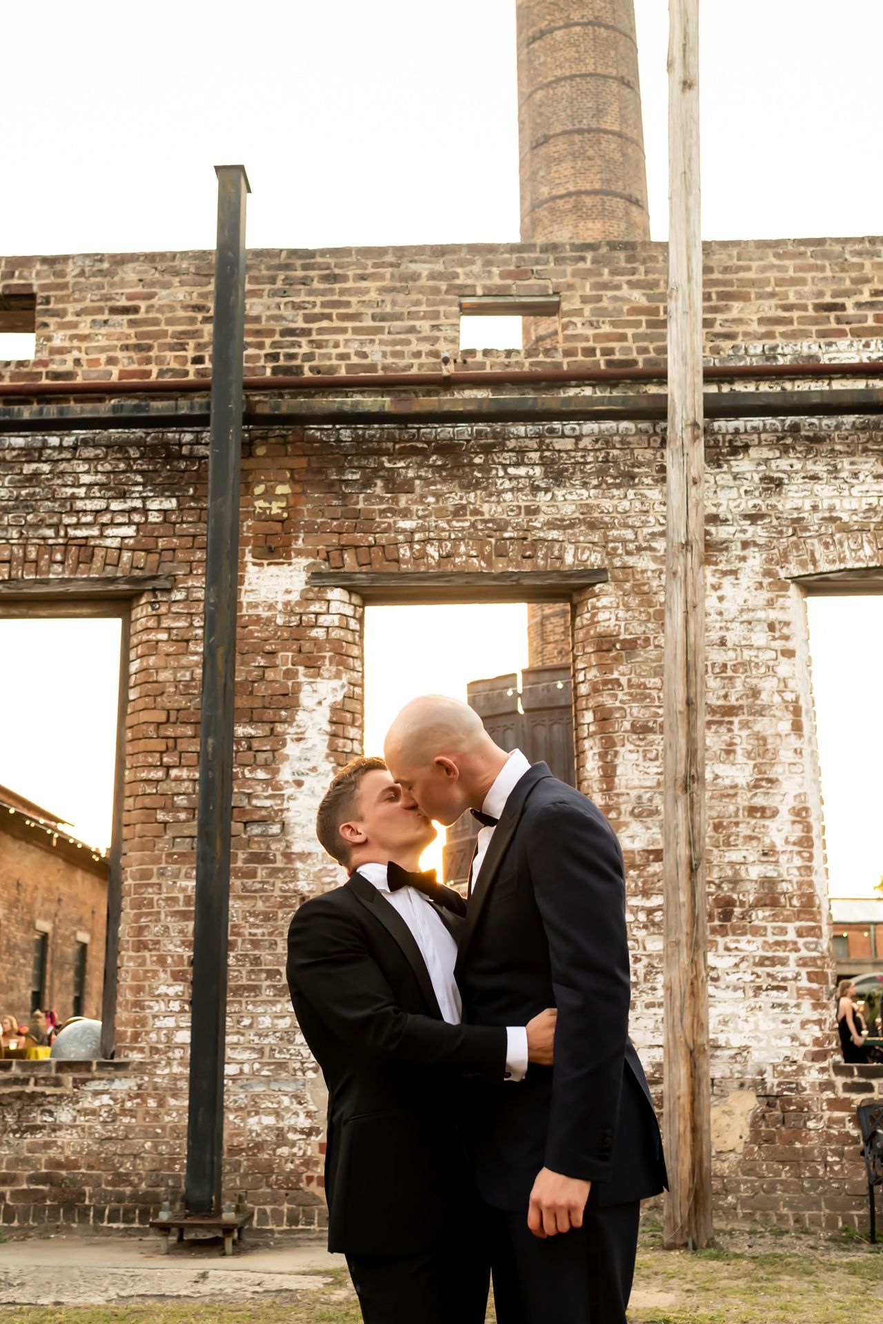 Two people in formal attire share a romantic moment against a rustic brick backdrop with sunlight filtering through.