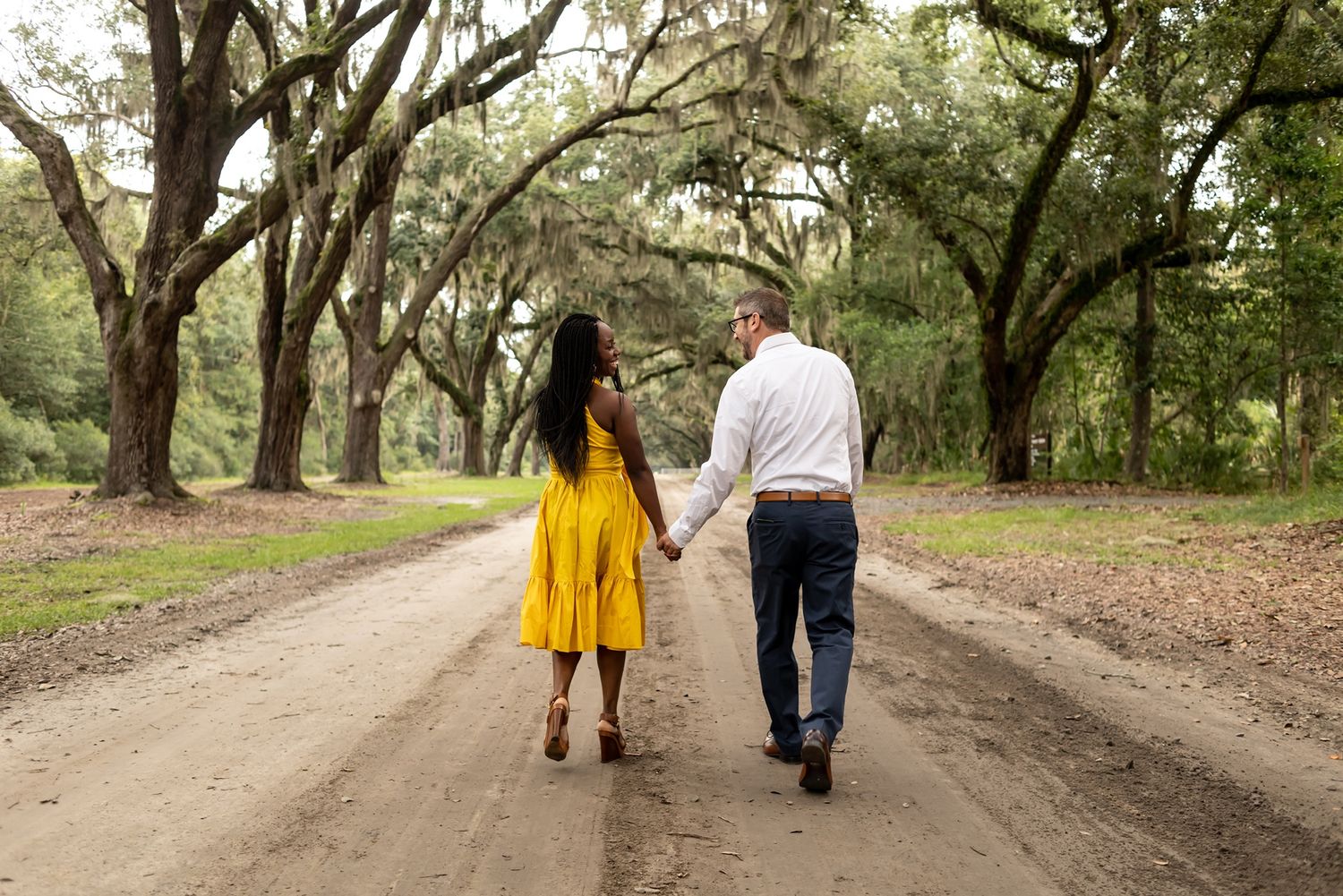 A couple walking together down a tree-lined dirt road, with one person wearing a bright yellow dress.