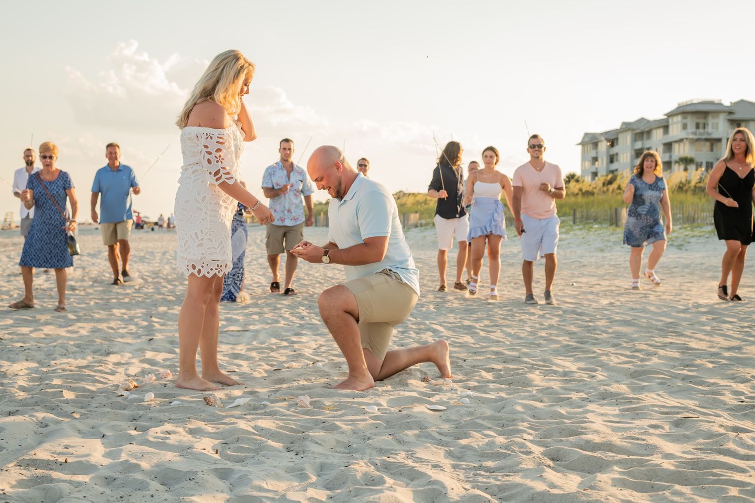 A romantic proposal moment unfolds on a sandy beach at sunset as onlookers gather to witness the special occasion.