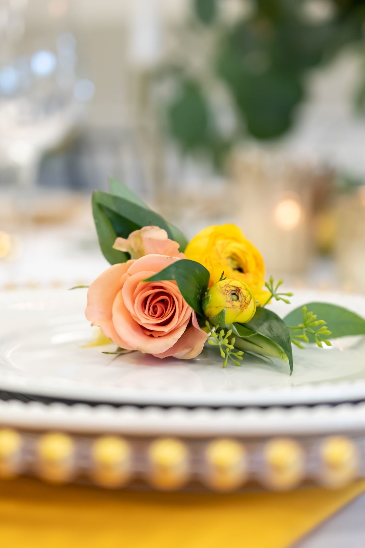 A pink rose and yellow ranunculus flower arrangement sits atop a white plate on a yellow tablecloth with soft bokeh background.