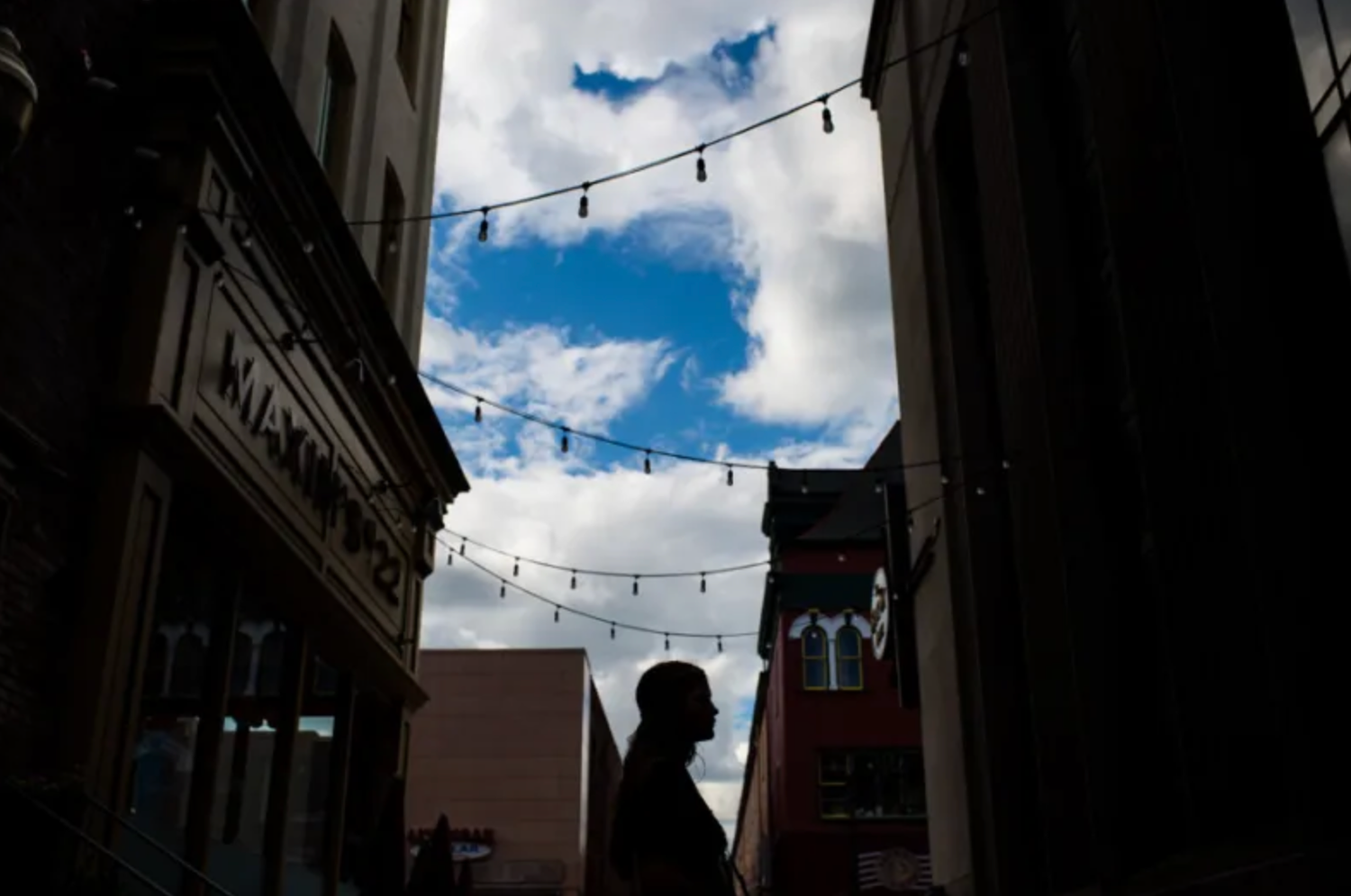 A silhouetted figure stands between buildings under string lights and blue cloudy sky in an urban setting.