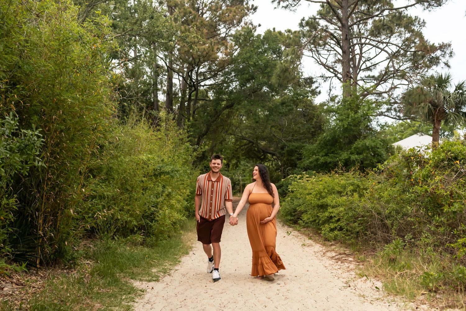 A couple walking along a sunny dirt path surrounded by lush green vegetation and tall trees.