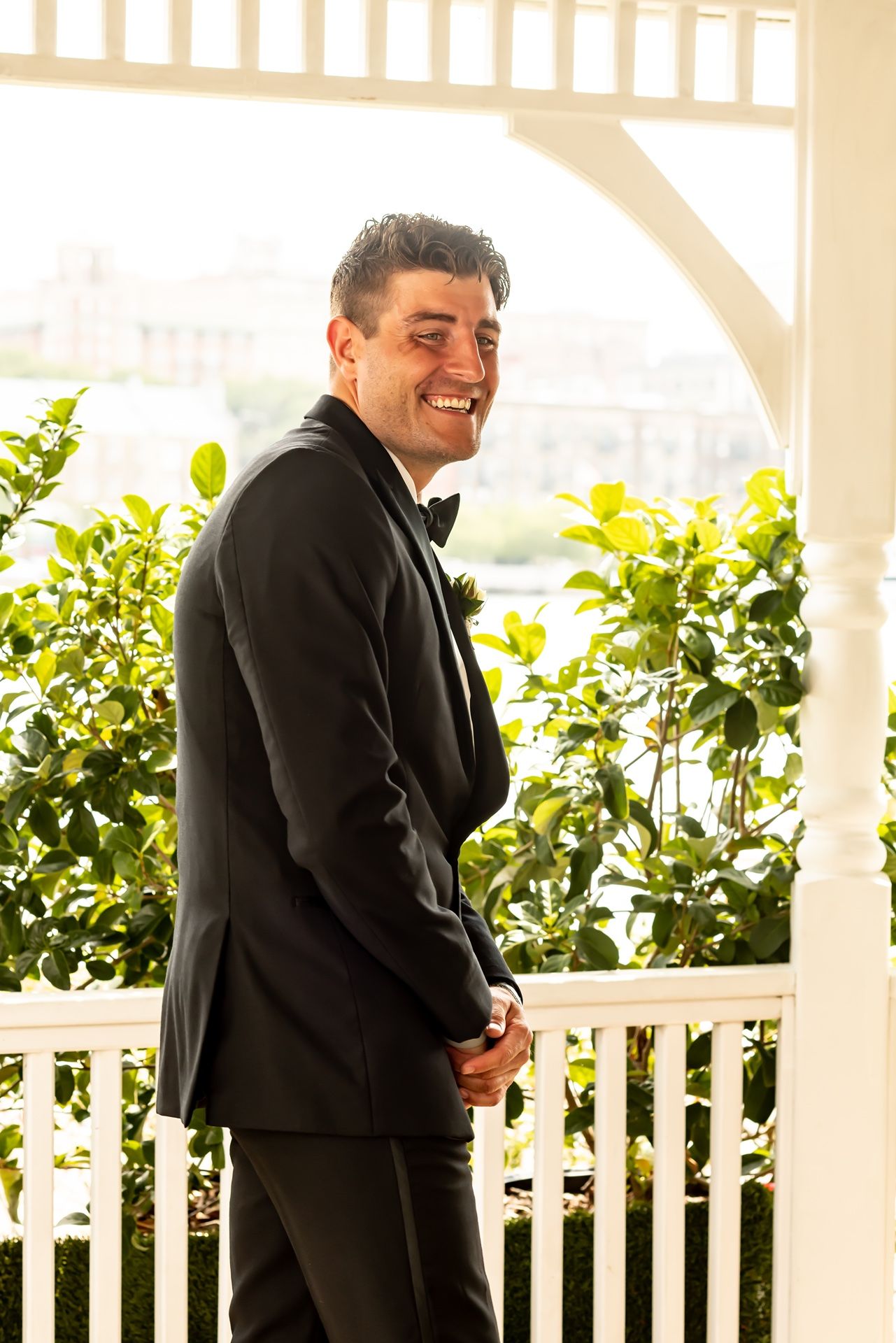 A person in a dark suit stands smiling on a white porch with lush greenery and sunlit scenery in the background.