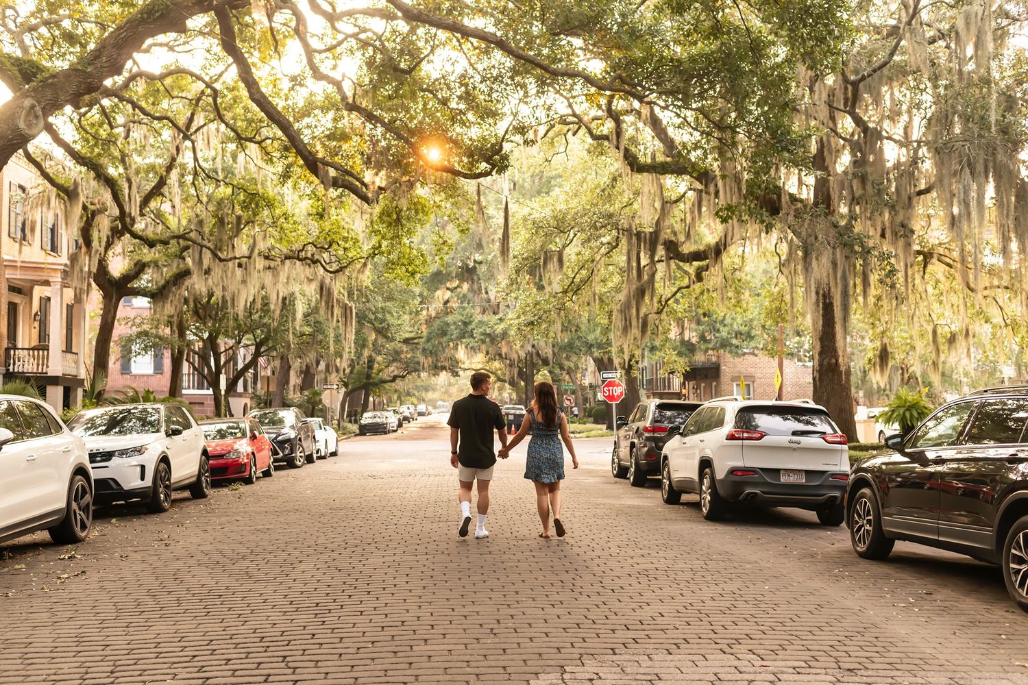 Two people walking down a historic Savannah street lined with Spanish moss-draped oak trees and parked cars.