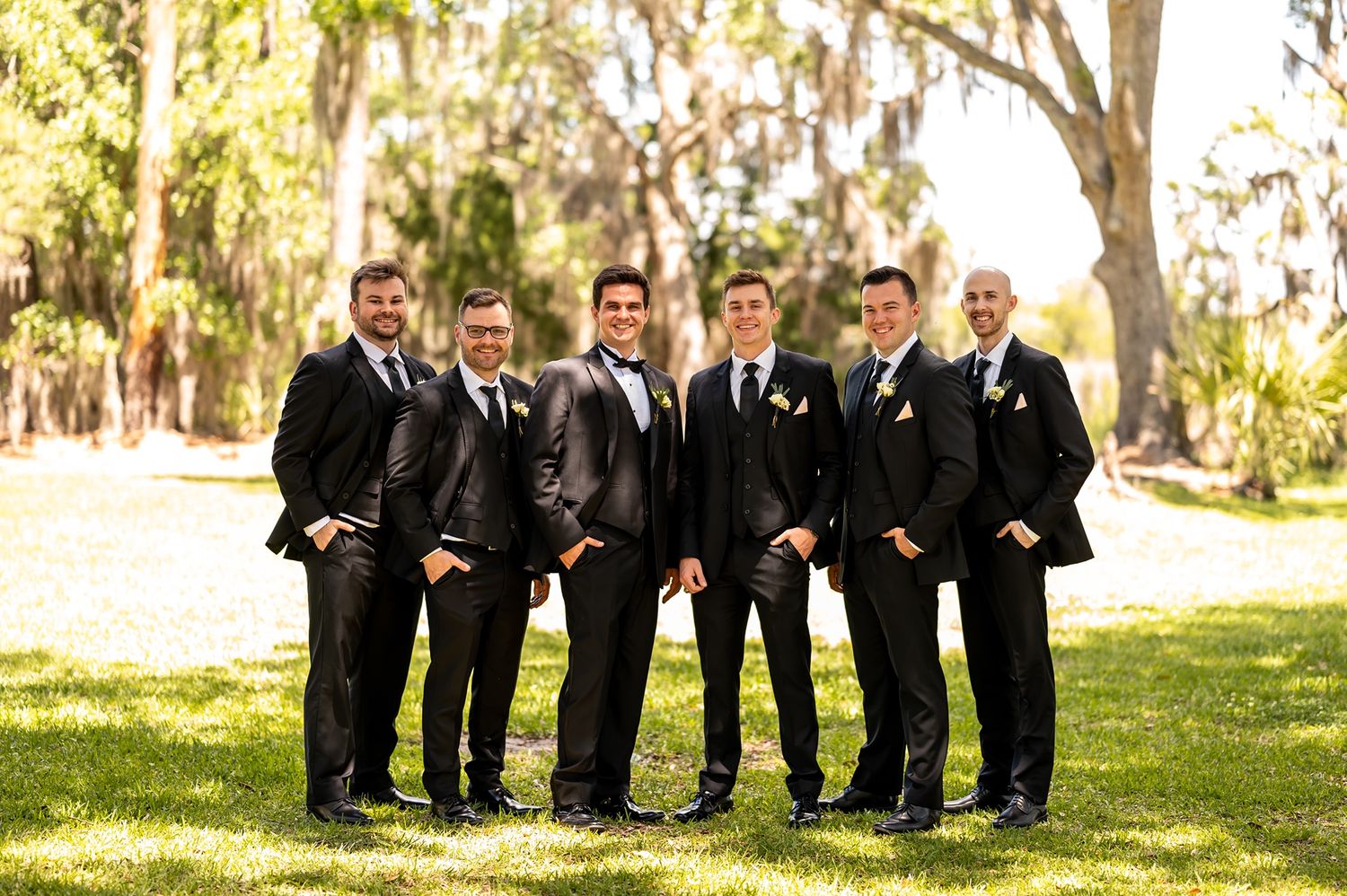 Groomsmen in black suits sit on grass near trees while posing for a casual wedding photo portrait.