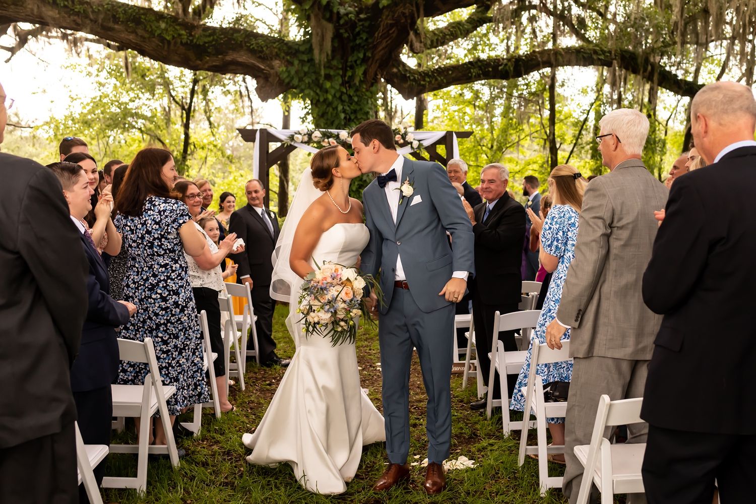 A newly married couple shares their first kiss at an outdoor wedding ceremony surrounded by guests and Spanish moss trees.
