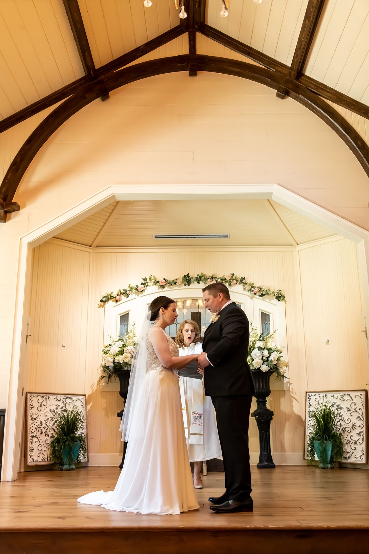 An intimate wedding ceremony takes place on a wooden stage under vaulted ceilings with floral arch decorations.