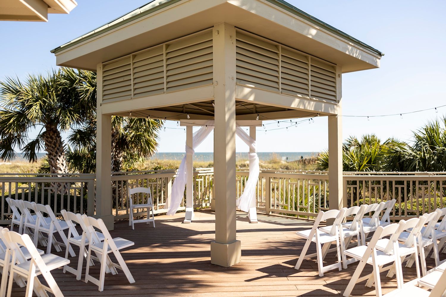 White beachfront wedding gazebo with wooden deck and folding chairs arranged on a coastal boardwalk near palm trees.