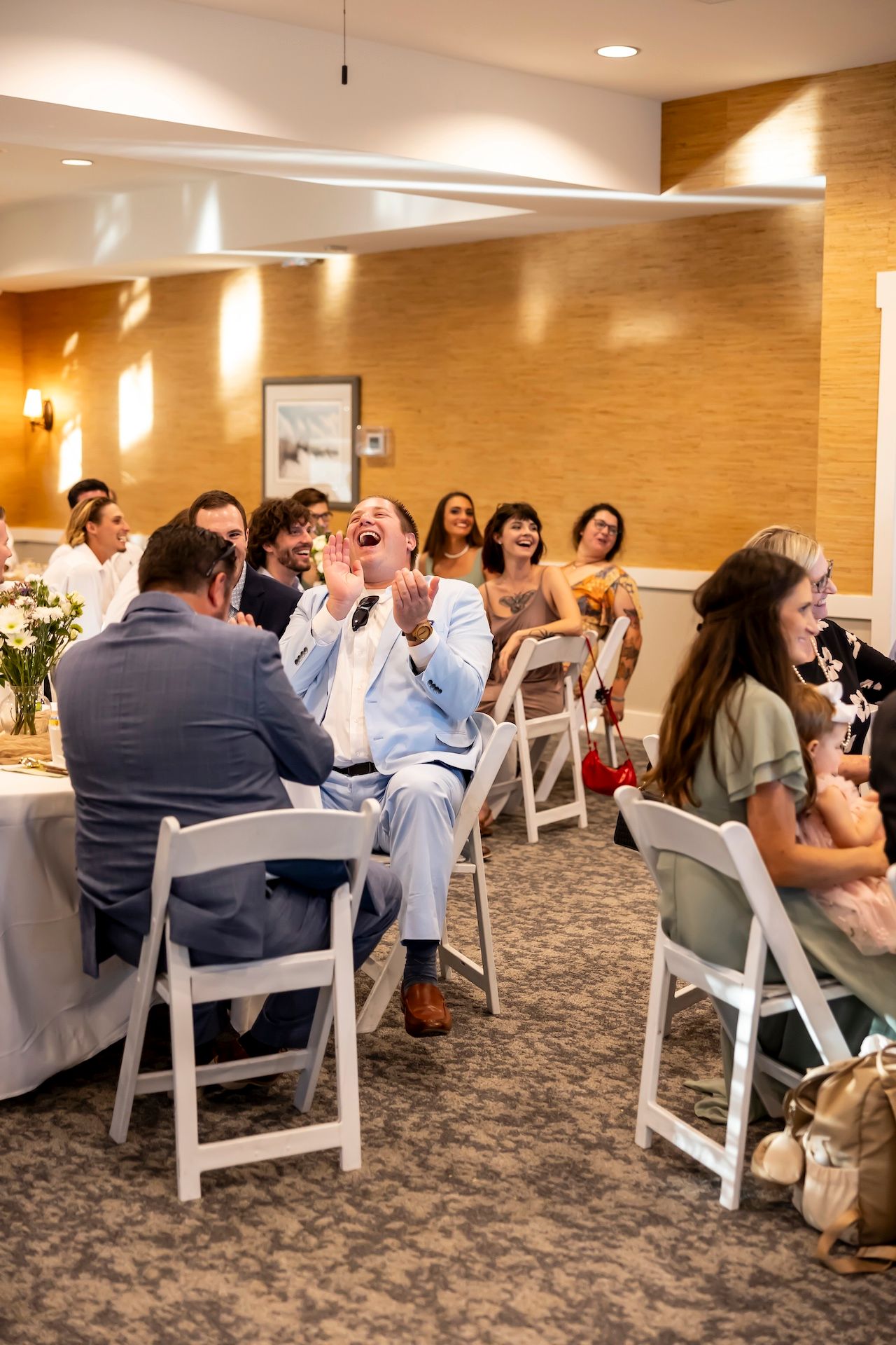 Wedding reception guests sit at white chairs in a banquet hall with warm wood paneled walls and recessed lighting.