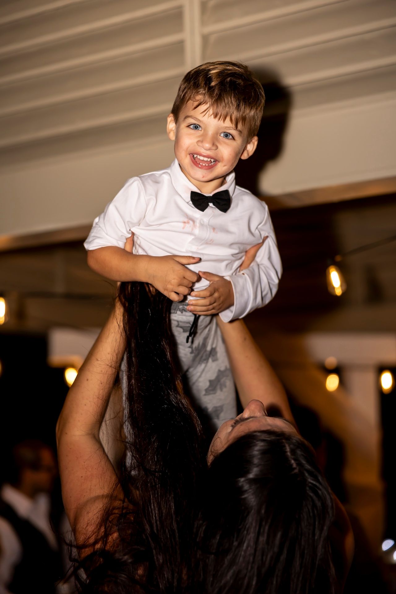 A young child wearing a white dress shirt and bowtie is lifted up joyfully at what appears to be a formal celebration.