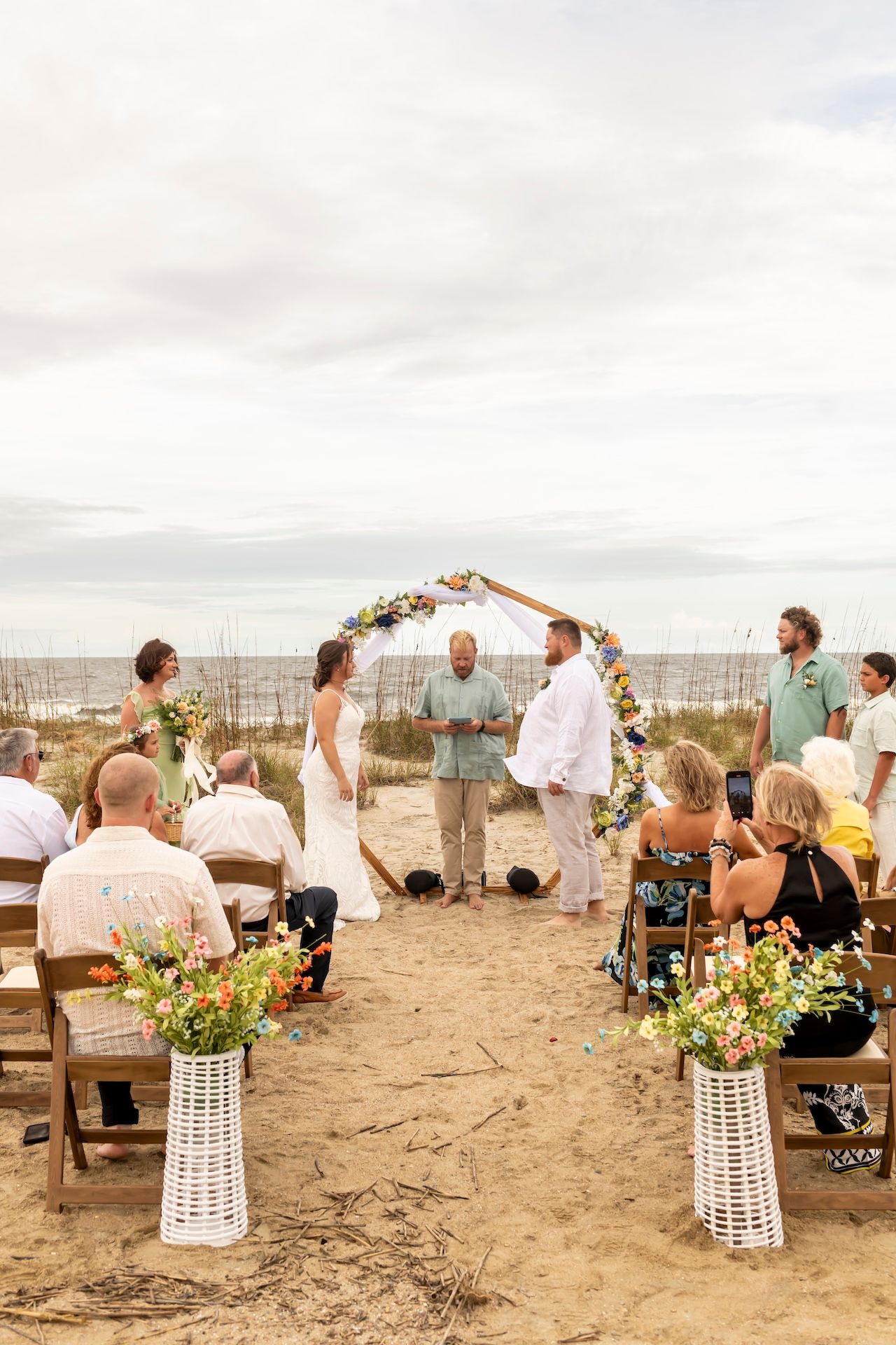 Outdoor beach wedding ceremony with circular floral arch and wooden chairs arranged on sandy ground overlooking the ocean.
