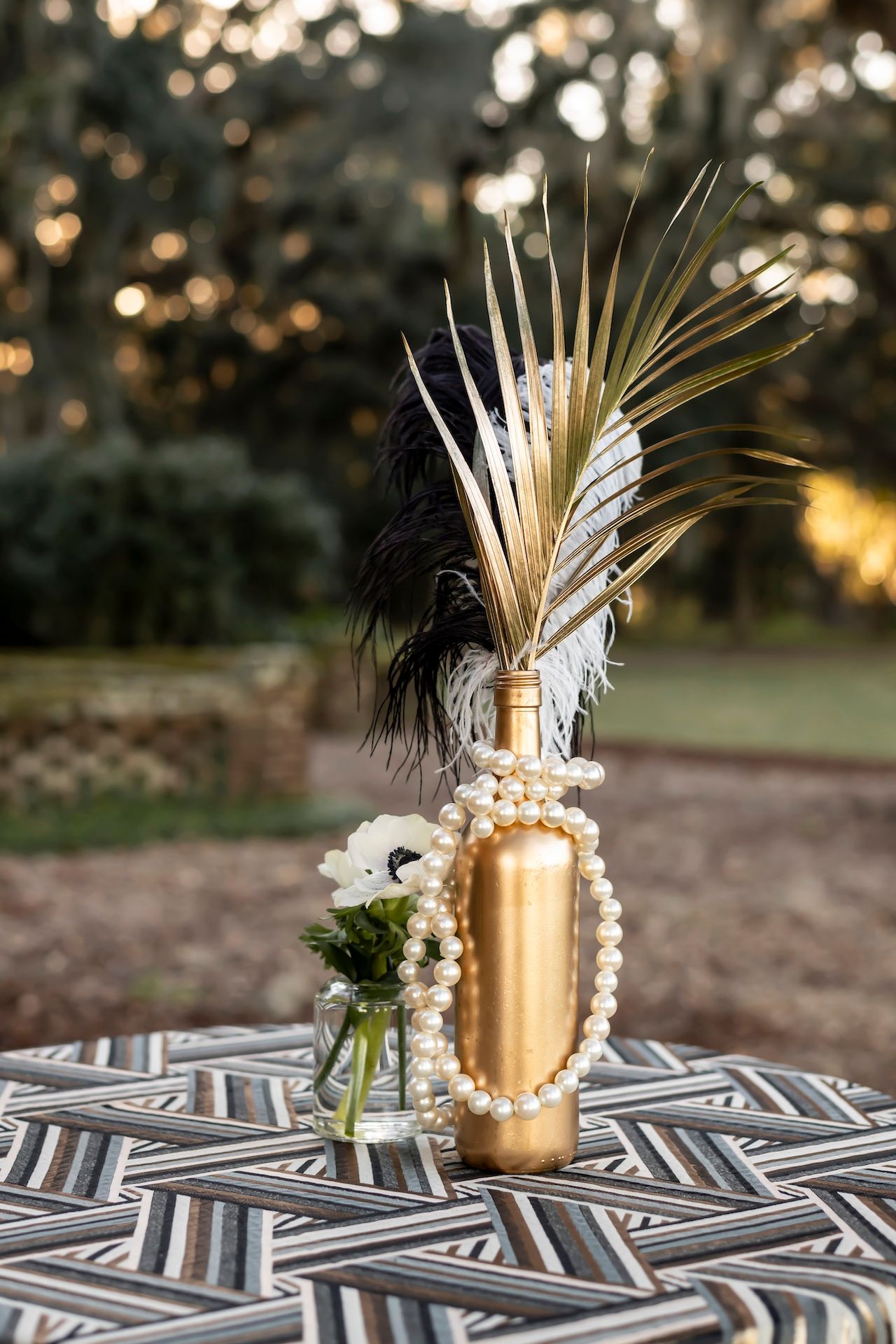 A gold painted wine bottle decorated with pearls and feathers sits on a geometric patterned tablecloth outdoors at sunset.
