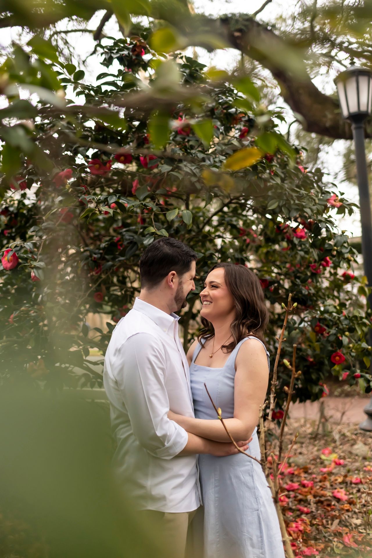 A couple embraces under a blooming camellia tree with a vintage lamp post in the background on an autumn day.