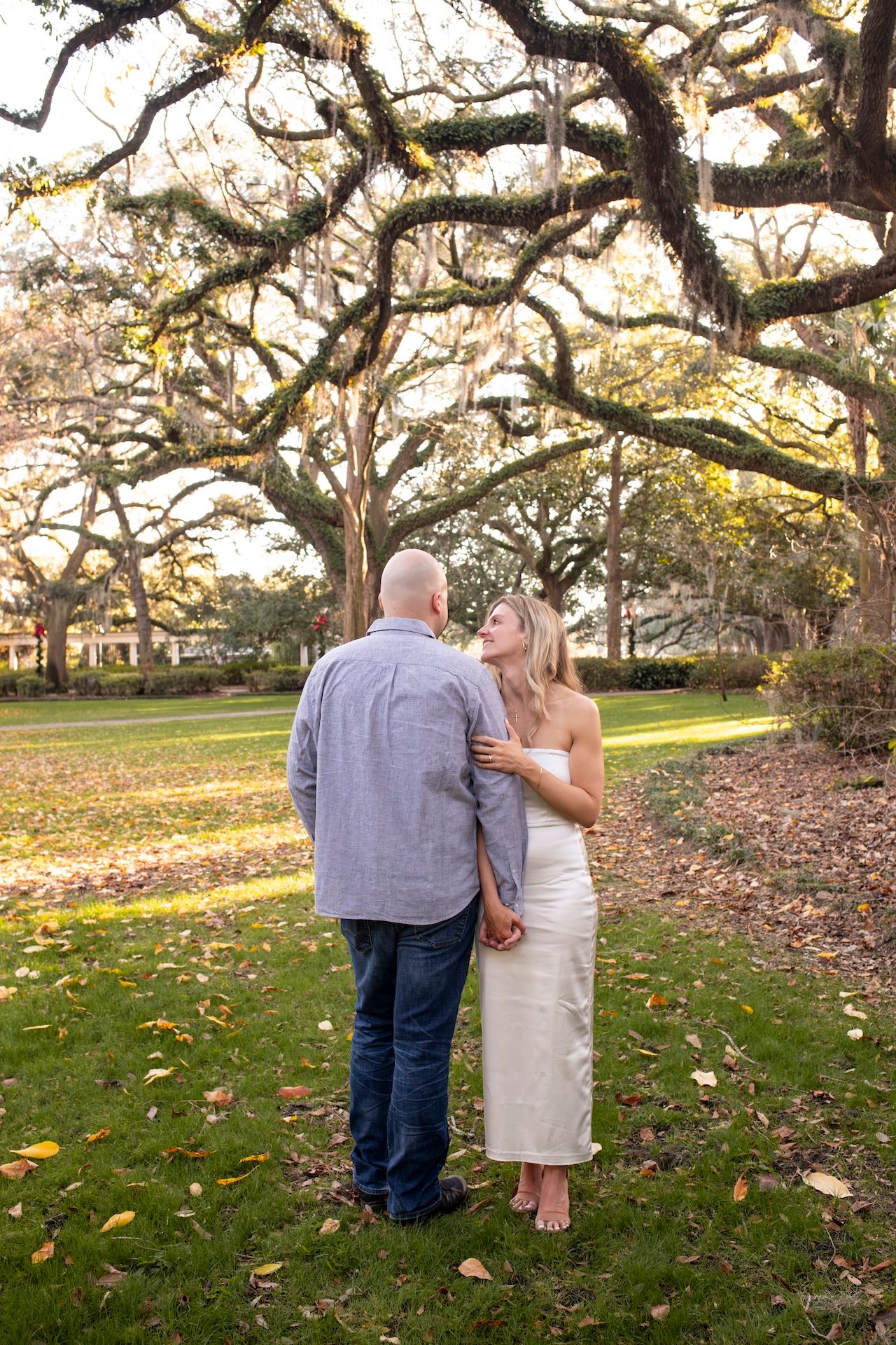 A couple shares an intimate moment beneath sprawling moss-covered oak trees on a grassy lawn during golden hour.