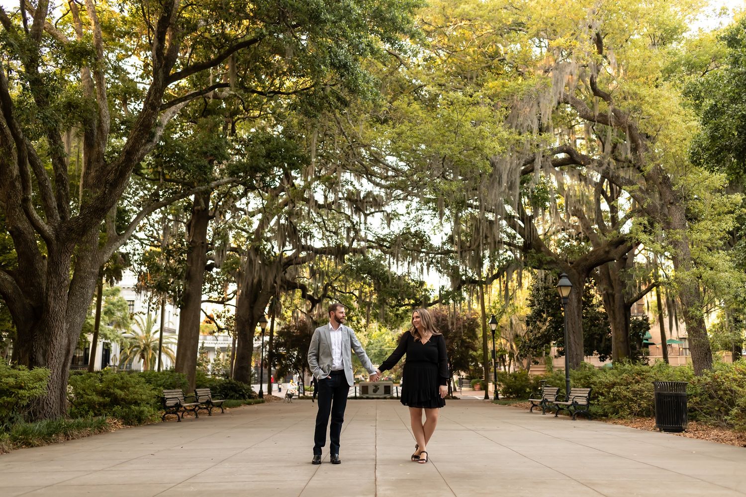 A couple walks together on a tree-lined path with Spanish moss hanging from the branches above.