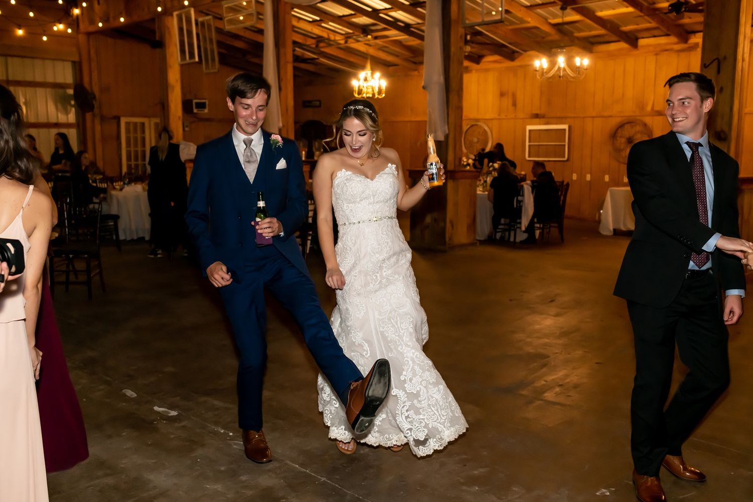 Wedding guests dance together at a rustic indoor venue with warm lighting and wooden beams overhead.