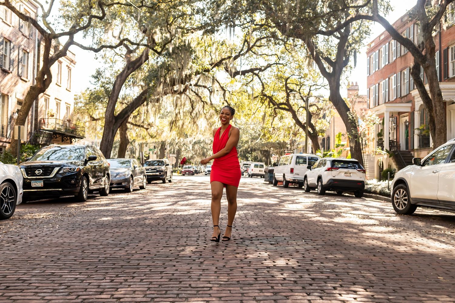 A scenic cobblestone street in Charleston lined with Spanish moss hanging from oak trees and parked cars on both sides.