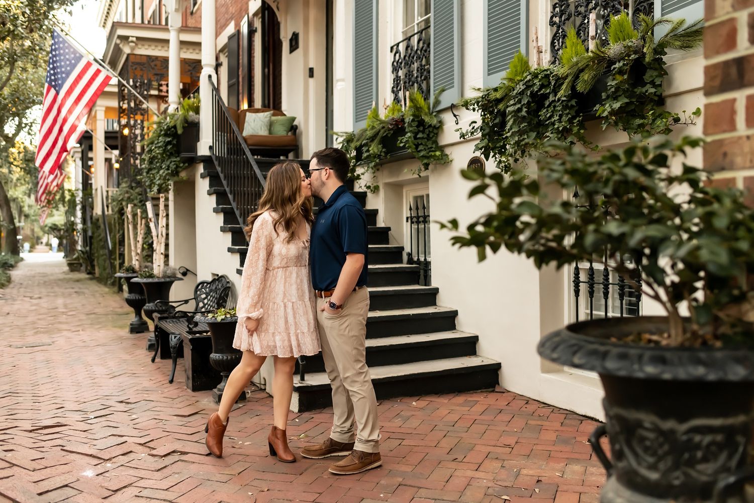 A romantic moment shared between two people on a charming cobblestone sidewalk with an American flag and greenery.