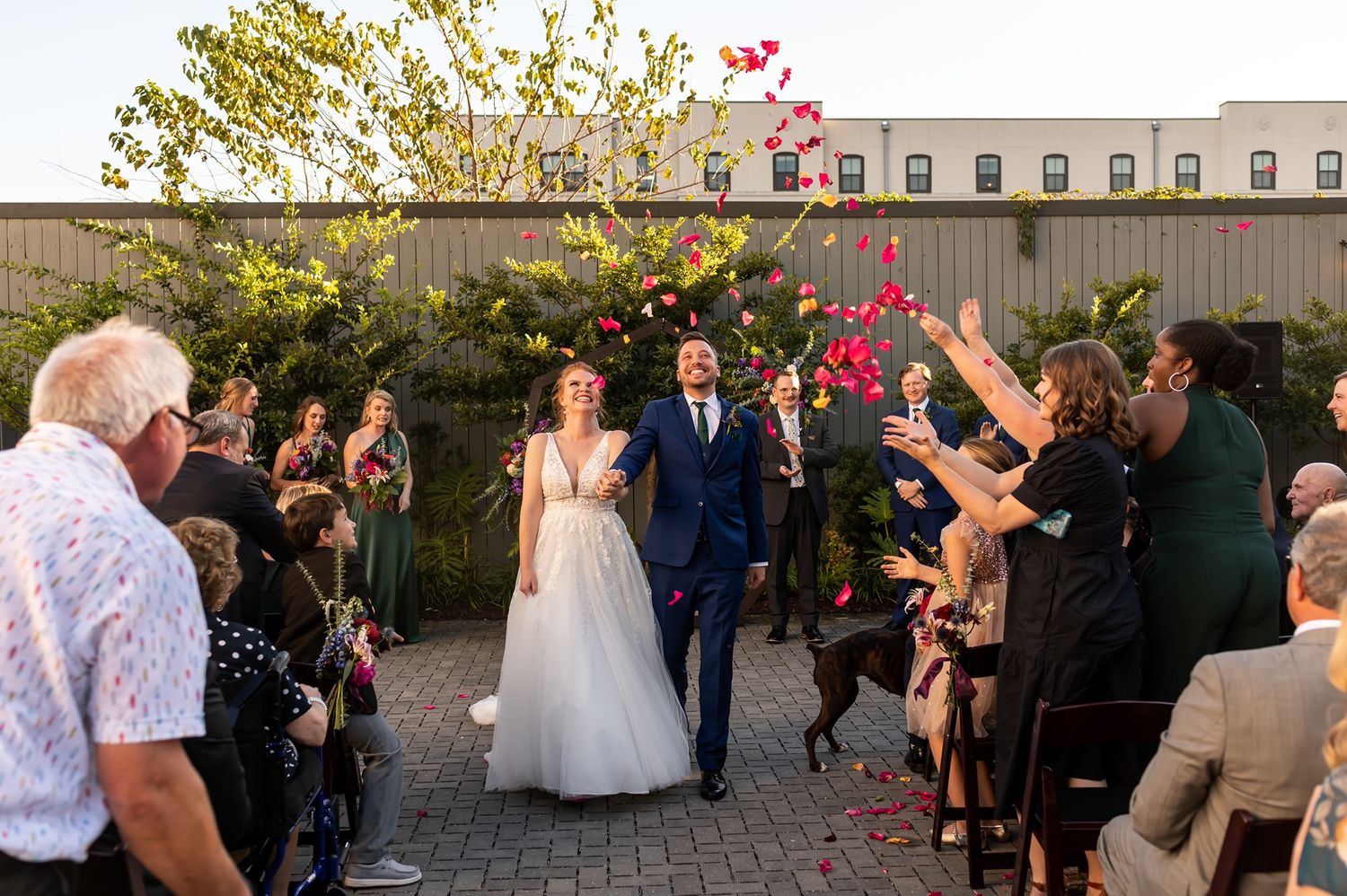 Wedding guests celebrate by throwing pink rose petals during an outdoor garden celebration at sunset.