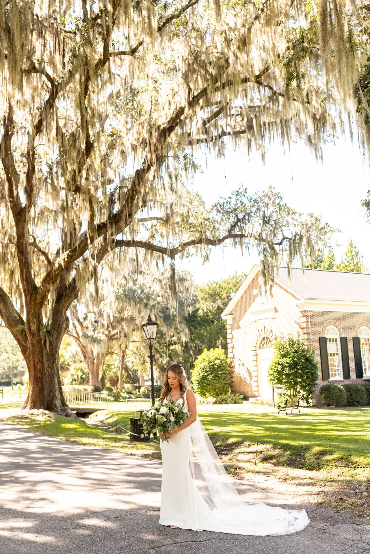 A bride in a flowing white gown poses beneath Spanish moss draped trees on a garden path near a charming house.