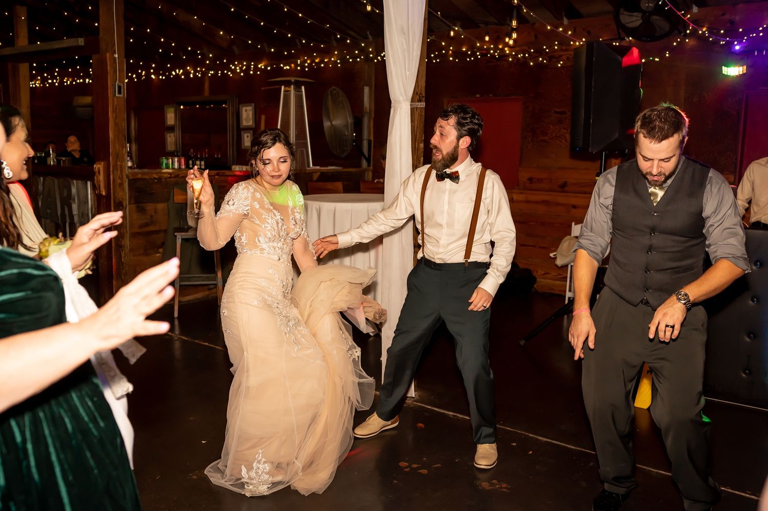 Wedding party dances energetically on the dance floor at a reception with decorative string lights overhead.