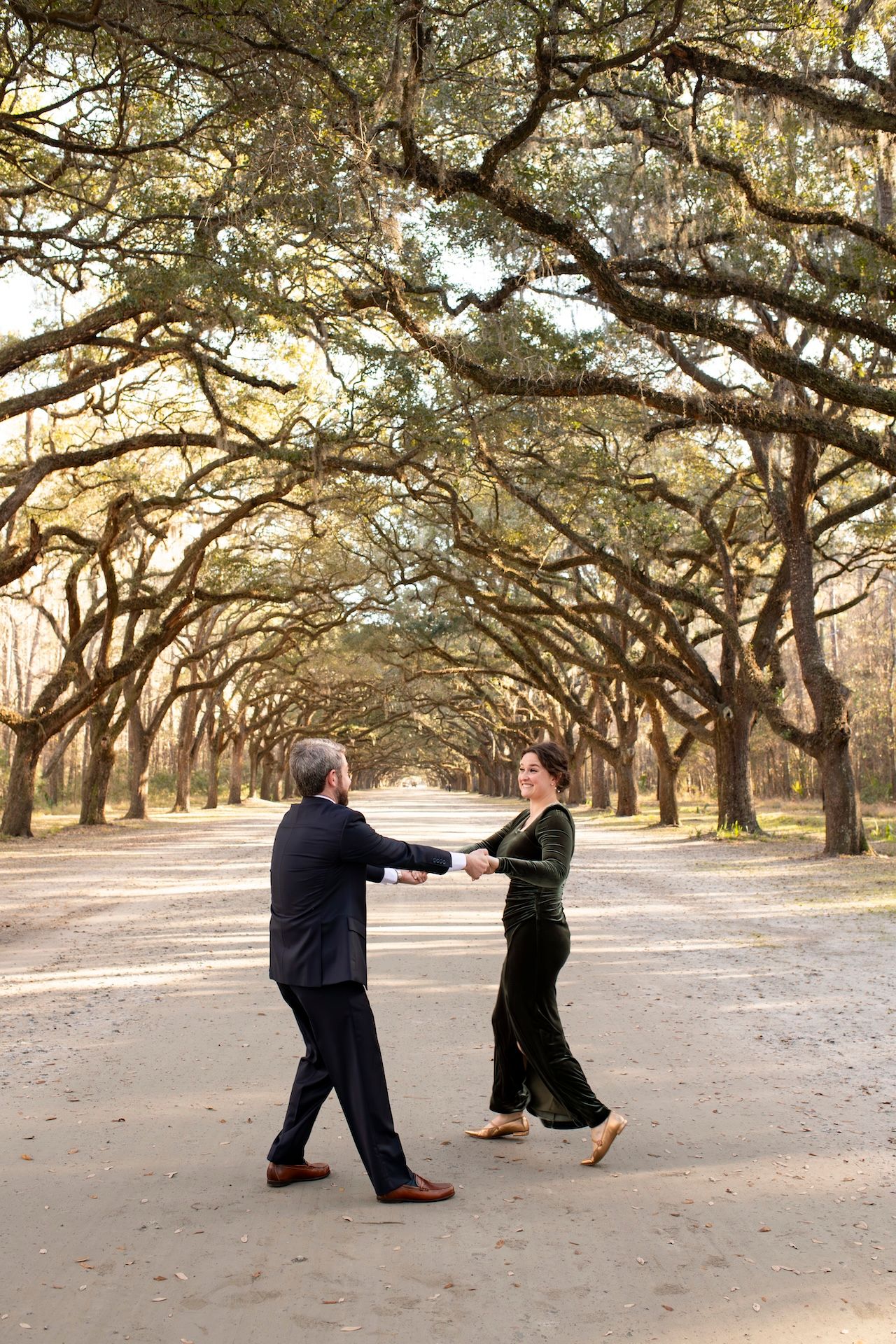 Two people dance together on a path beneath a stunning canopy of oak trees draped with Spanish moss at sunset.