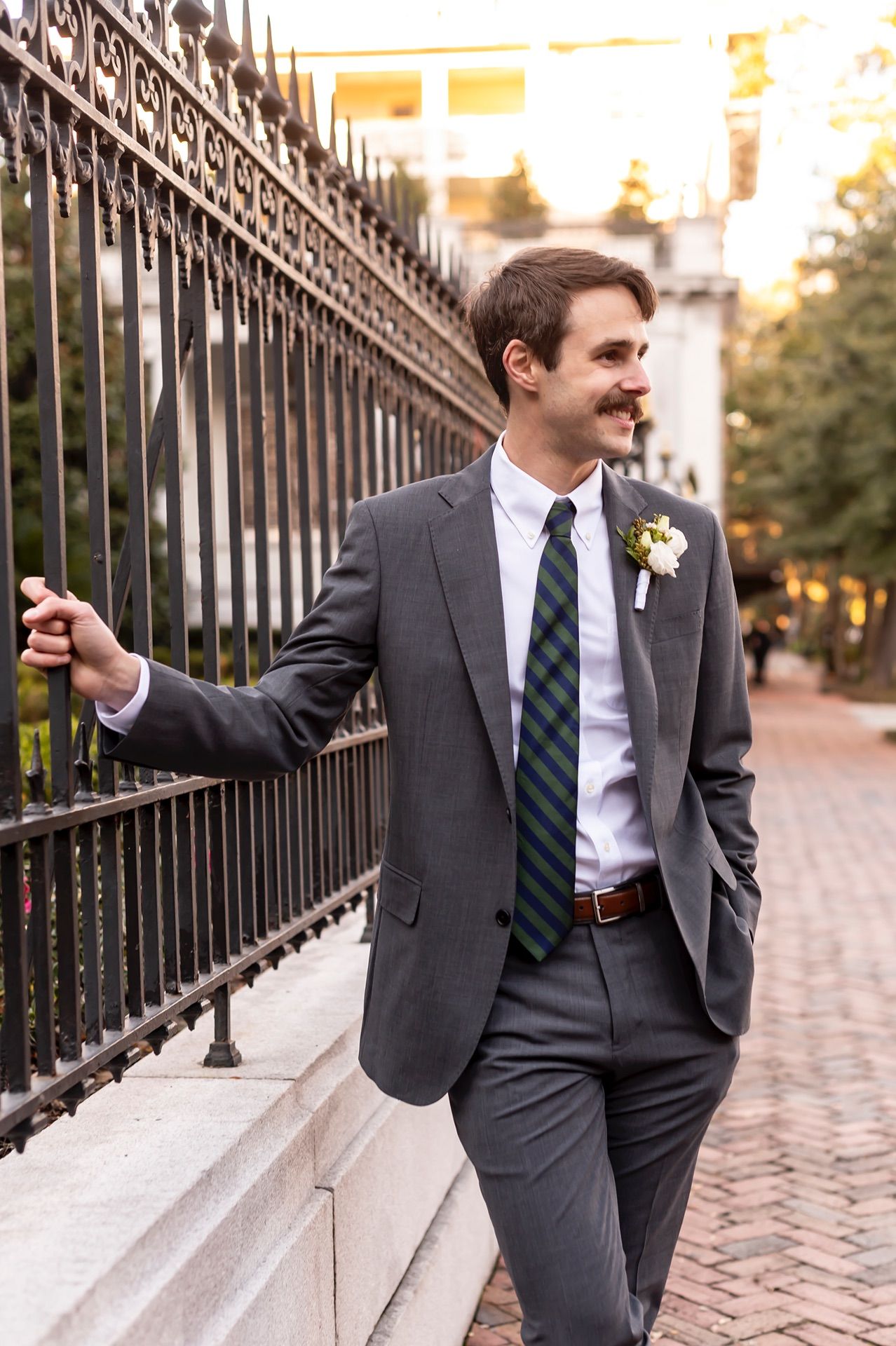 Candid shots of a groomsman in a dark suit and green tie posing along a decorative iron fence at sunset.