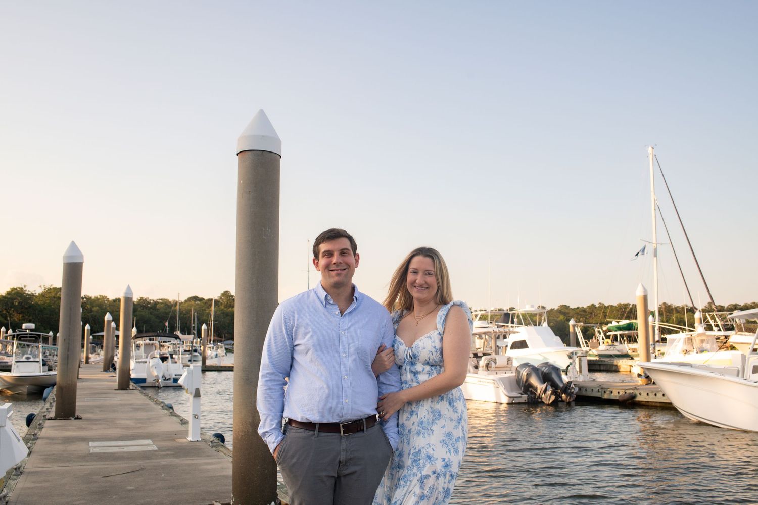 Couple poses for romantic photos at sunset by a marina with boats and water in the background.