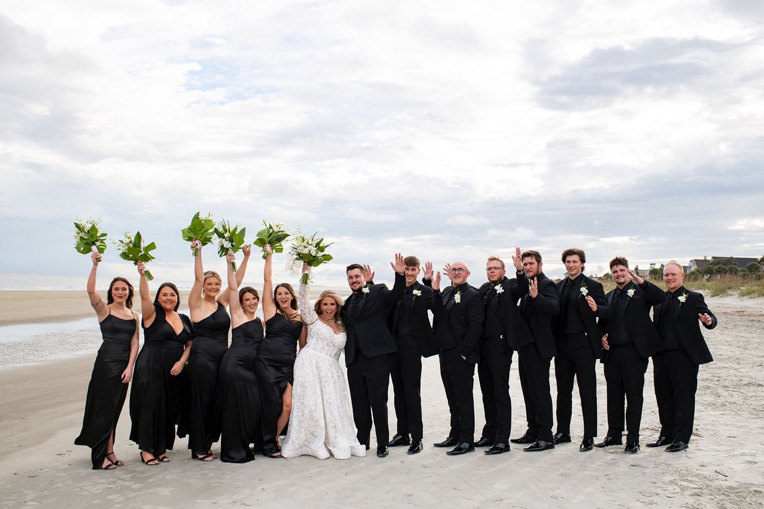 Wedding party in formal black and white attire poses together on a scenic beach at sunset.
