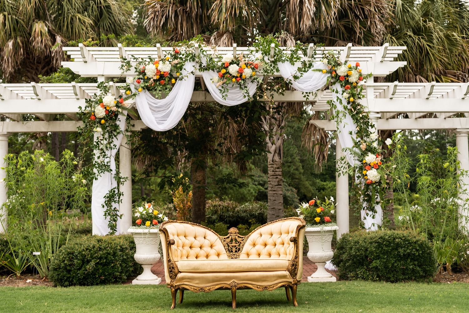 Elegant vintage sofa under white pergola decorated with floral arrangements and draping in a garden setting.