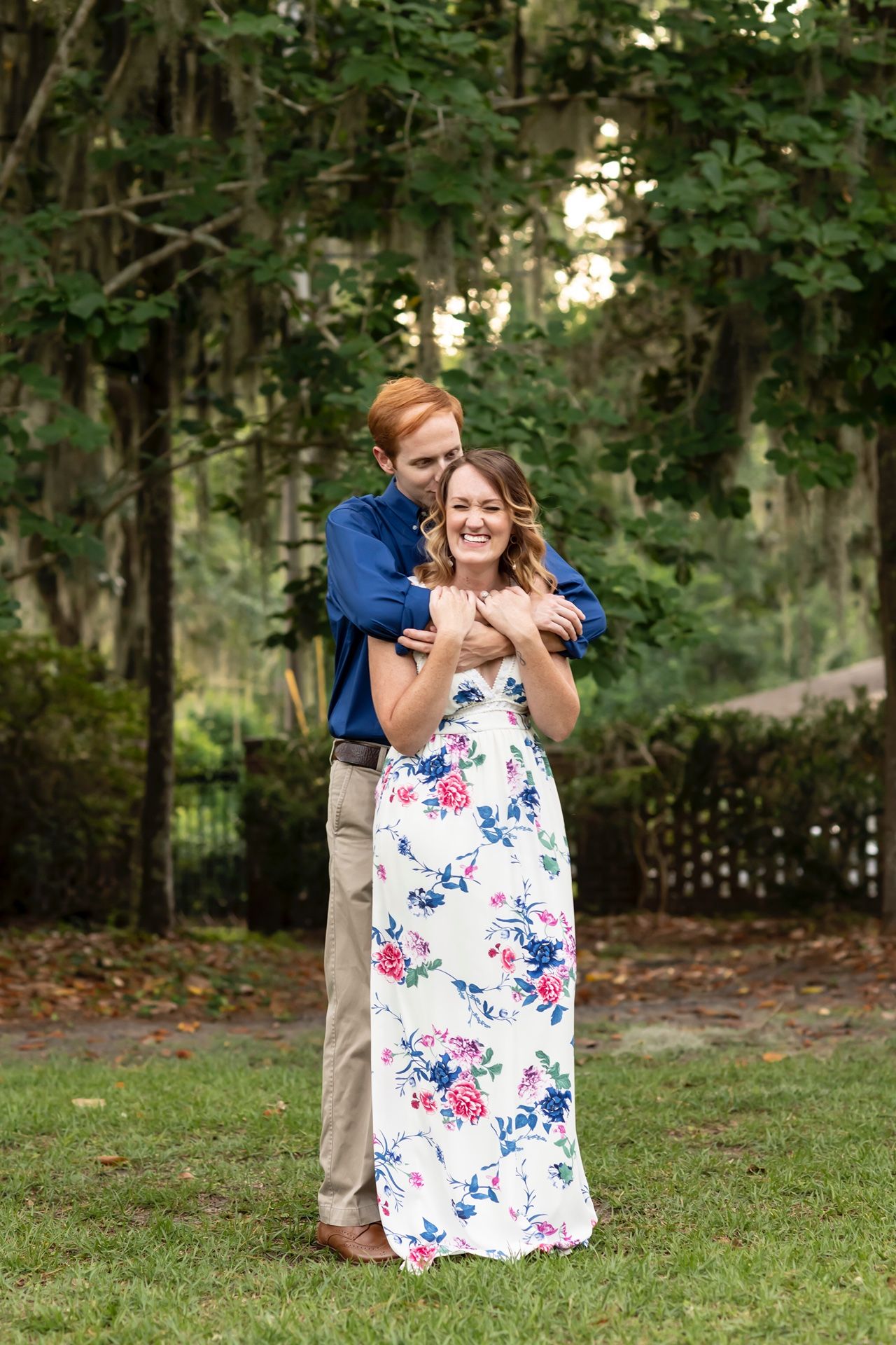 A couple embraces outdoors in a garden setting, with her wearing a white floral maxi dress and him in blue.