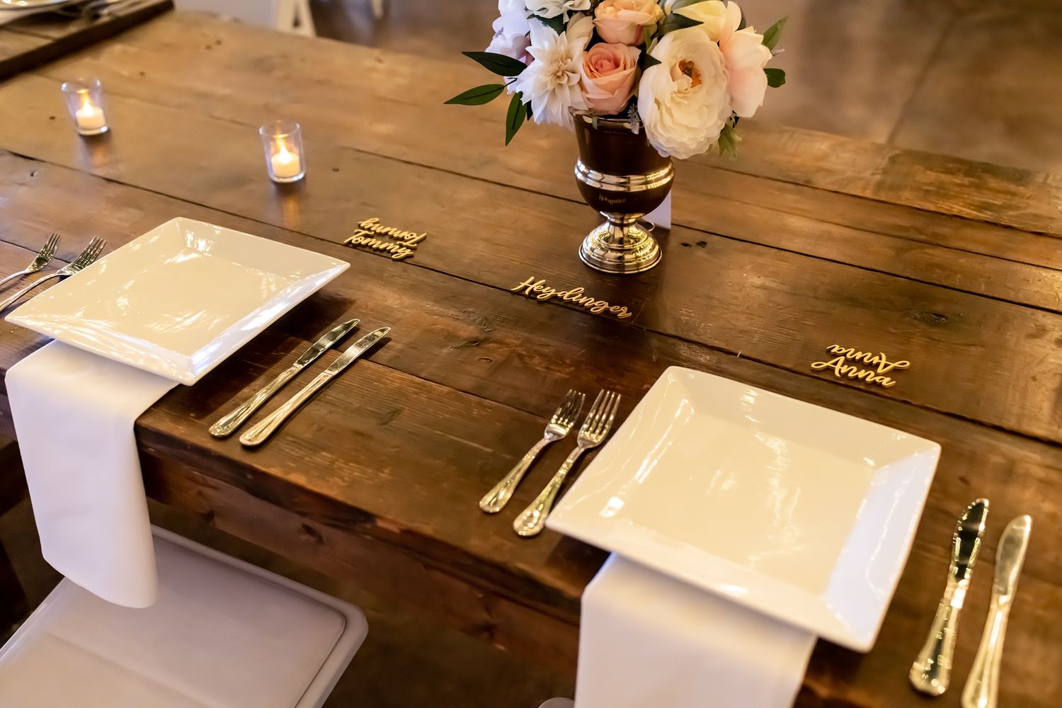 Elegant rustic table setting with white linens, silverware and floral centerpiece in gold vase on wooden farmhouse table.