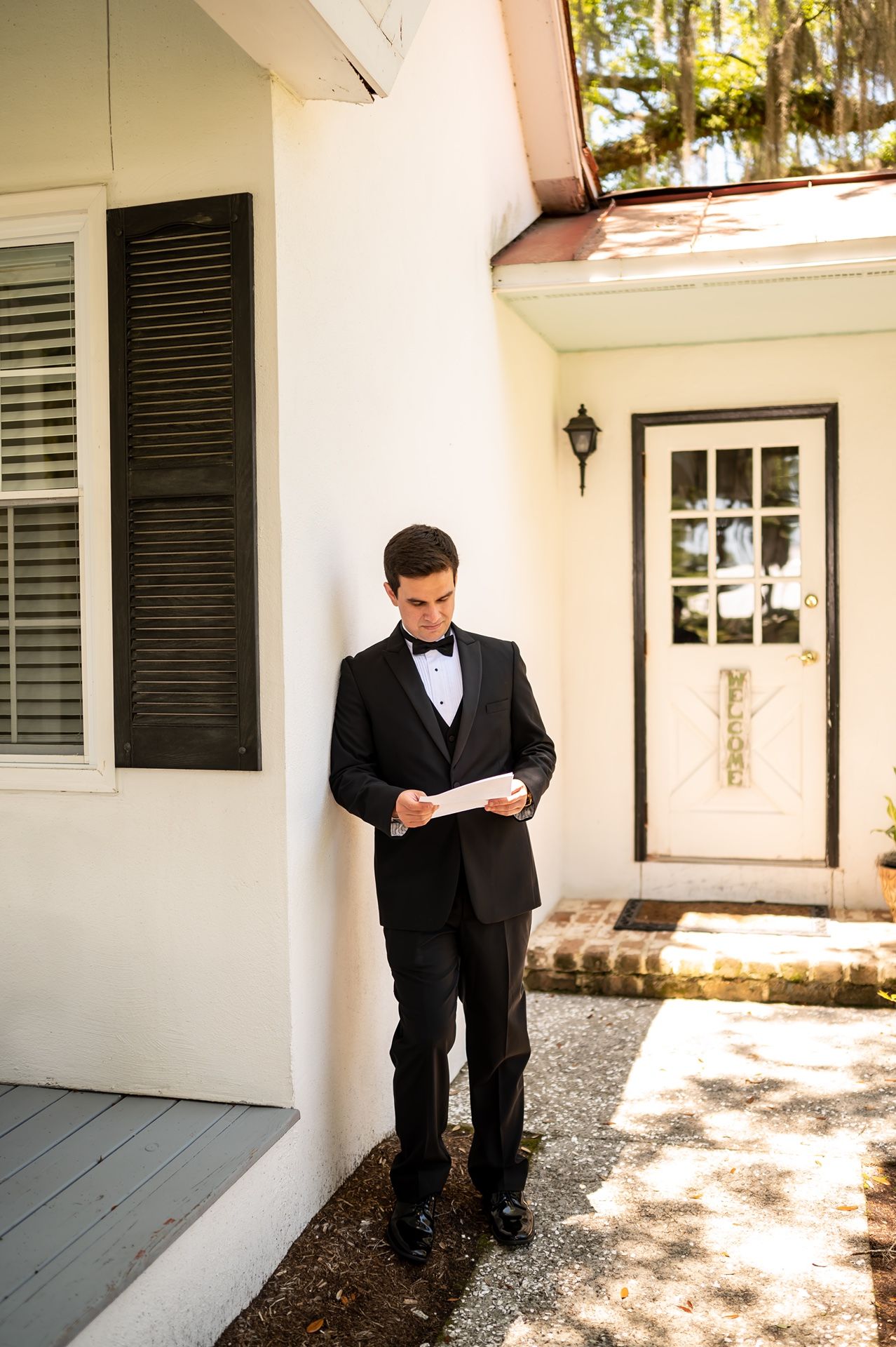 Person in formal black tuxedo reads letter while standing on a porch with white columns and black shutters.