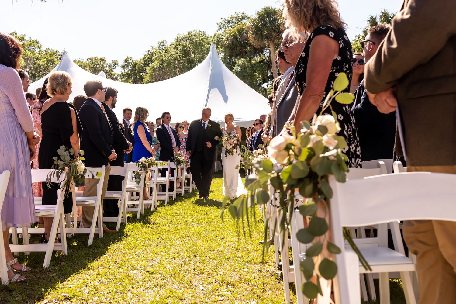 Outdoor wedding ceremony setup with white chairs decorated with greenery along aisle under tent on sunny day.