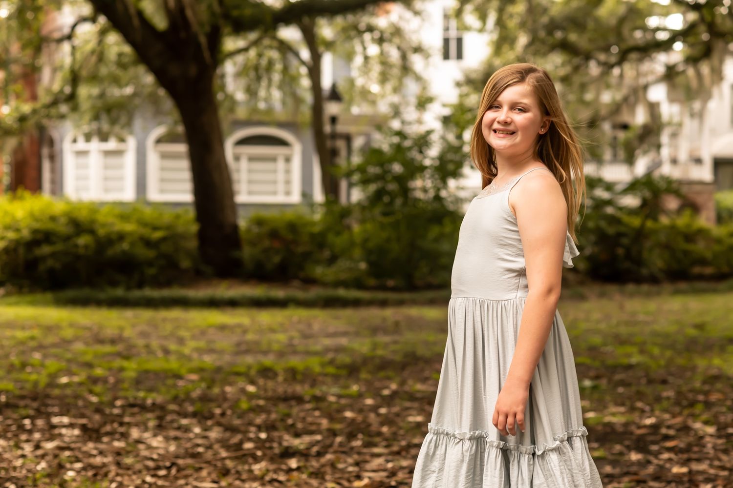 A person in a light tiered dress stands smiling in a garden setting with Spanish moss draped trees in the background.