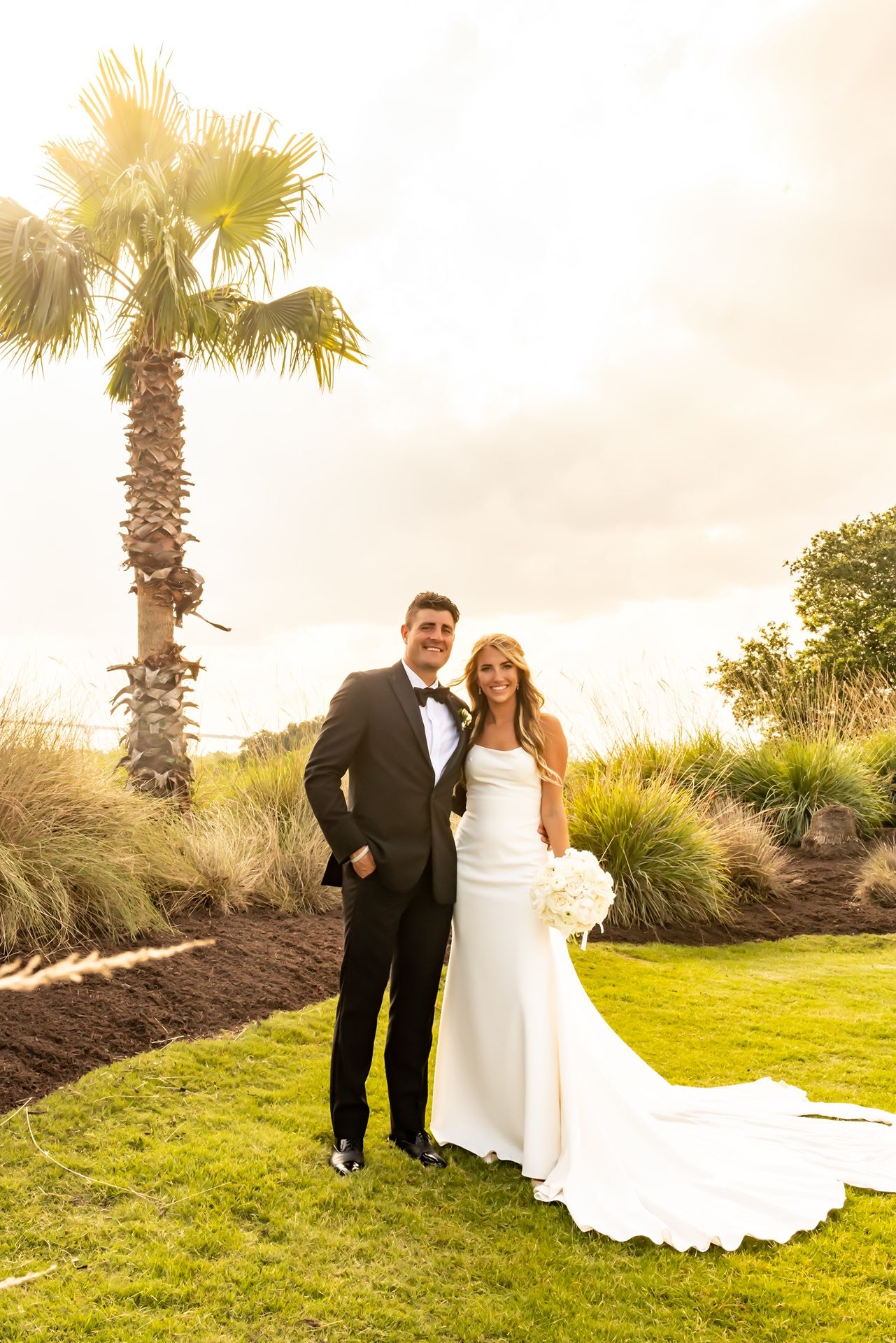 Wedding couple stands together on manicured lawn with palm tree and sunset sky in tropical resort setting.