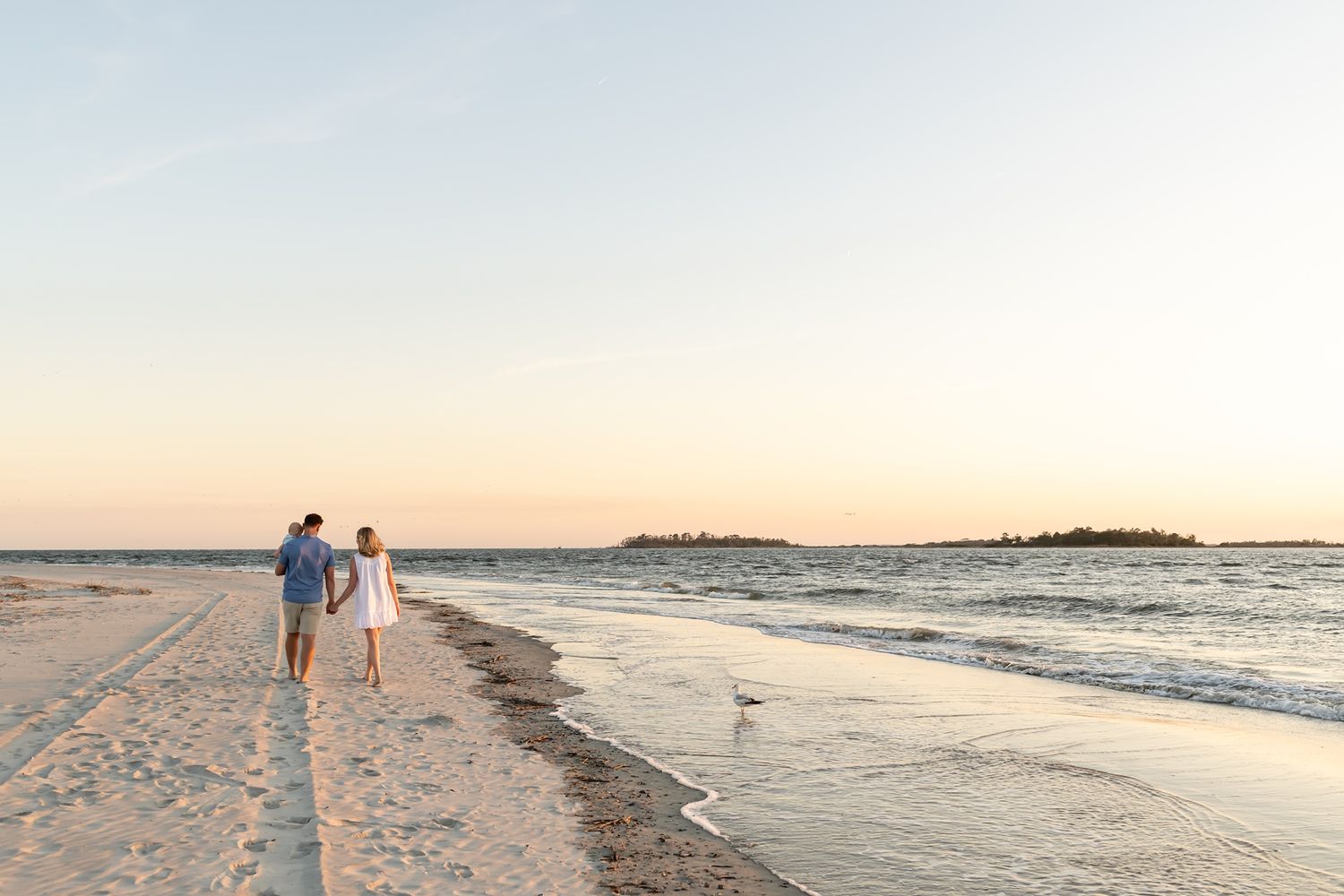 Two people walk hand in hand along a peaceful beach at sunset with golden light reflecting on the sand and water.