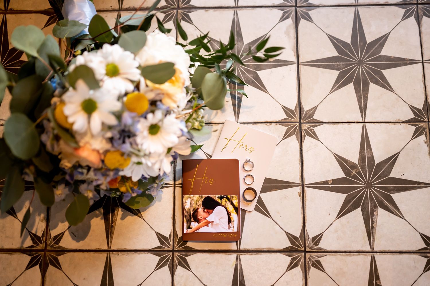 A wedding bouquet with white daisies and greenery rests on a star-patterned tile floor next to a vintage photo.