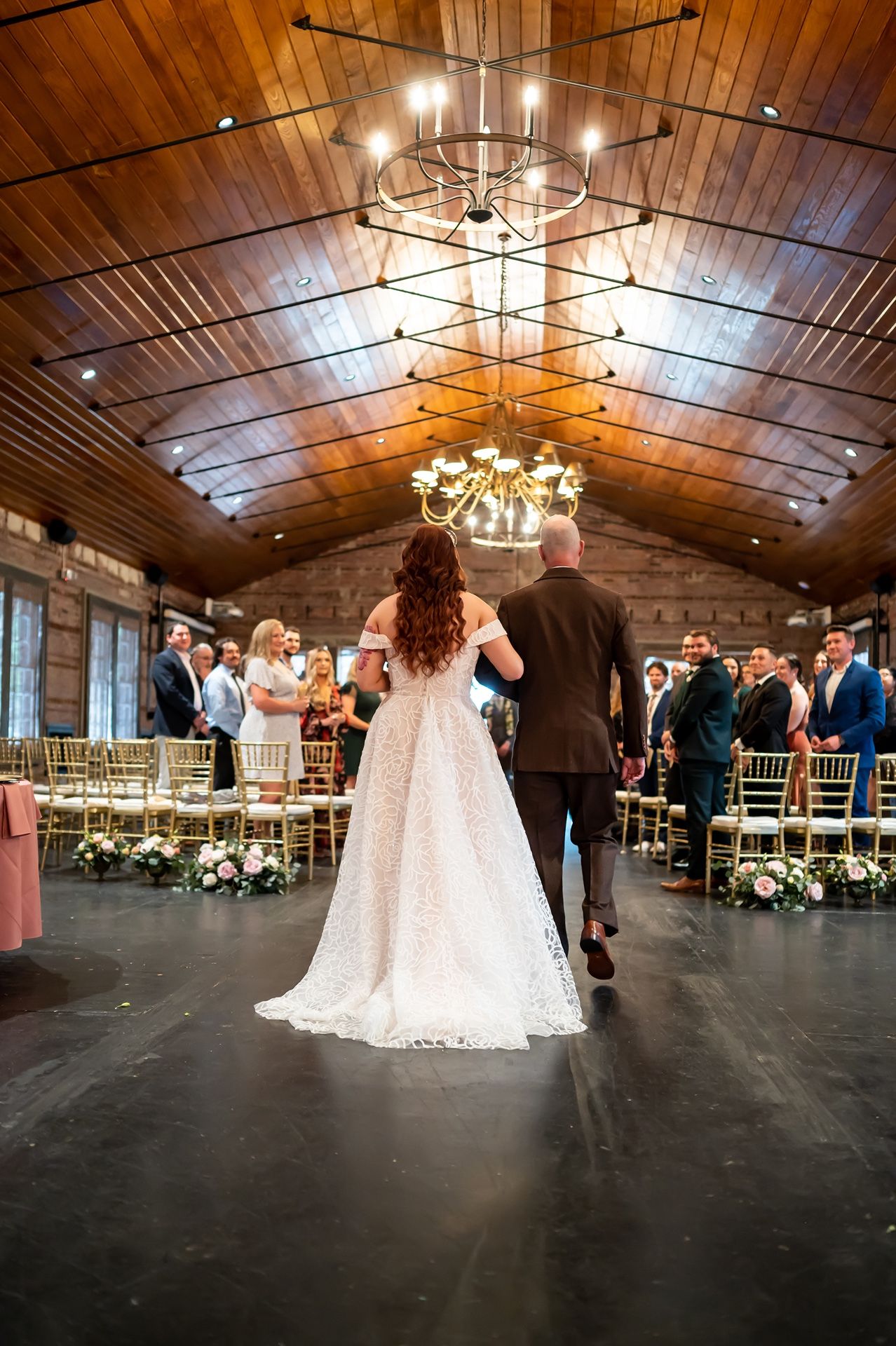 Couple enters rustic wedding venue with wooden vaulted ceiling, elegant chandeliers and floral arrangements lining the aisle.