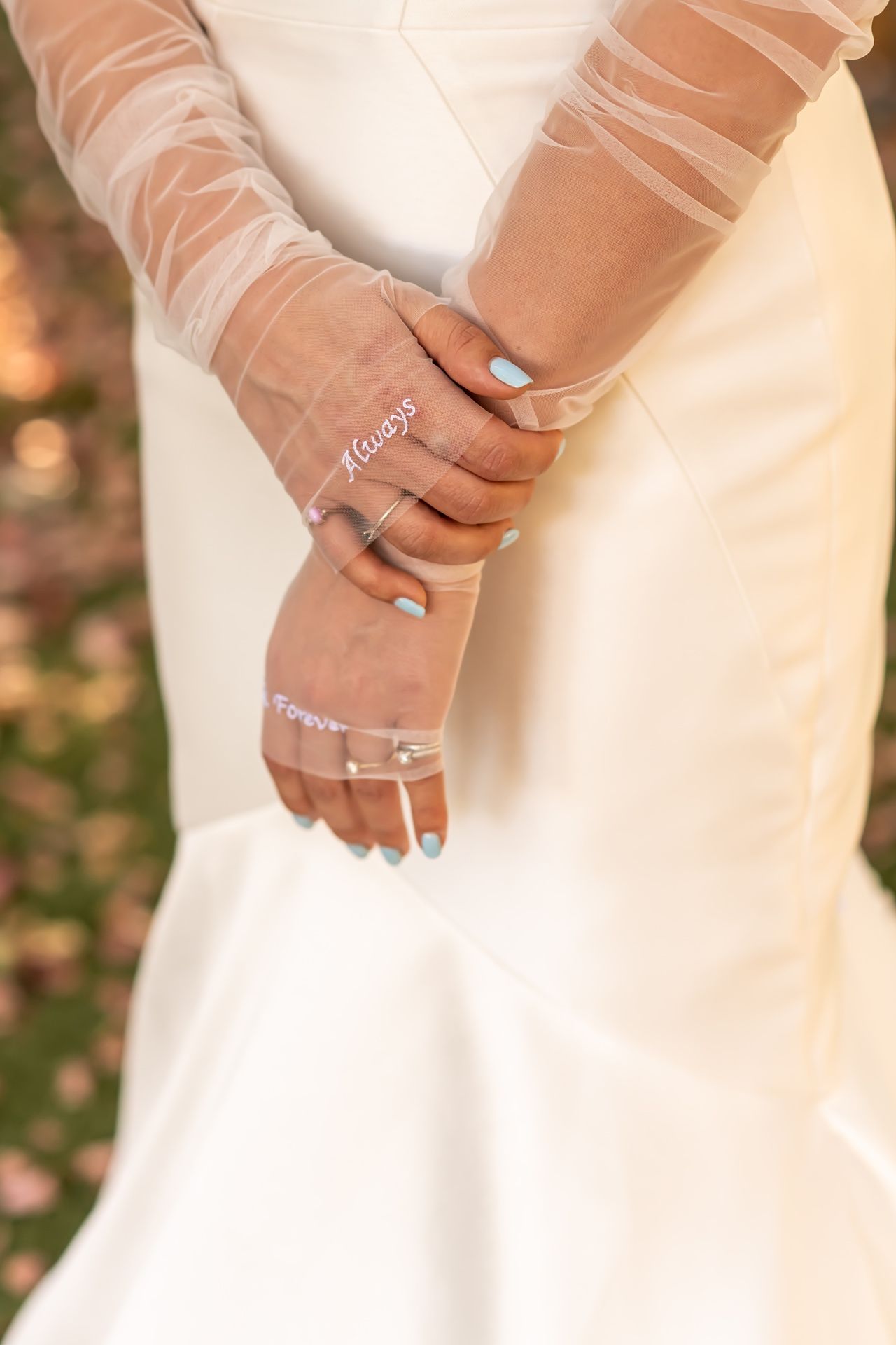 Hands wearing elegant sheer tulle bridal gloves with light blue nail polish and delicate jewelry against a white wedding dress.