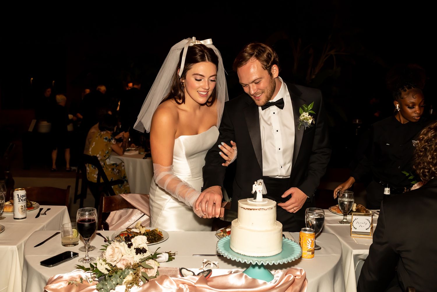 Bride and groom in formal attire jointly cut their white wedding cake at the reception dining table.