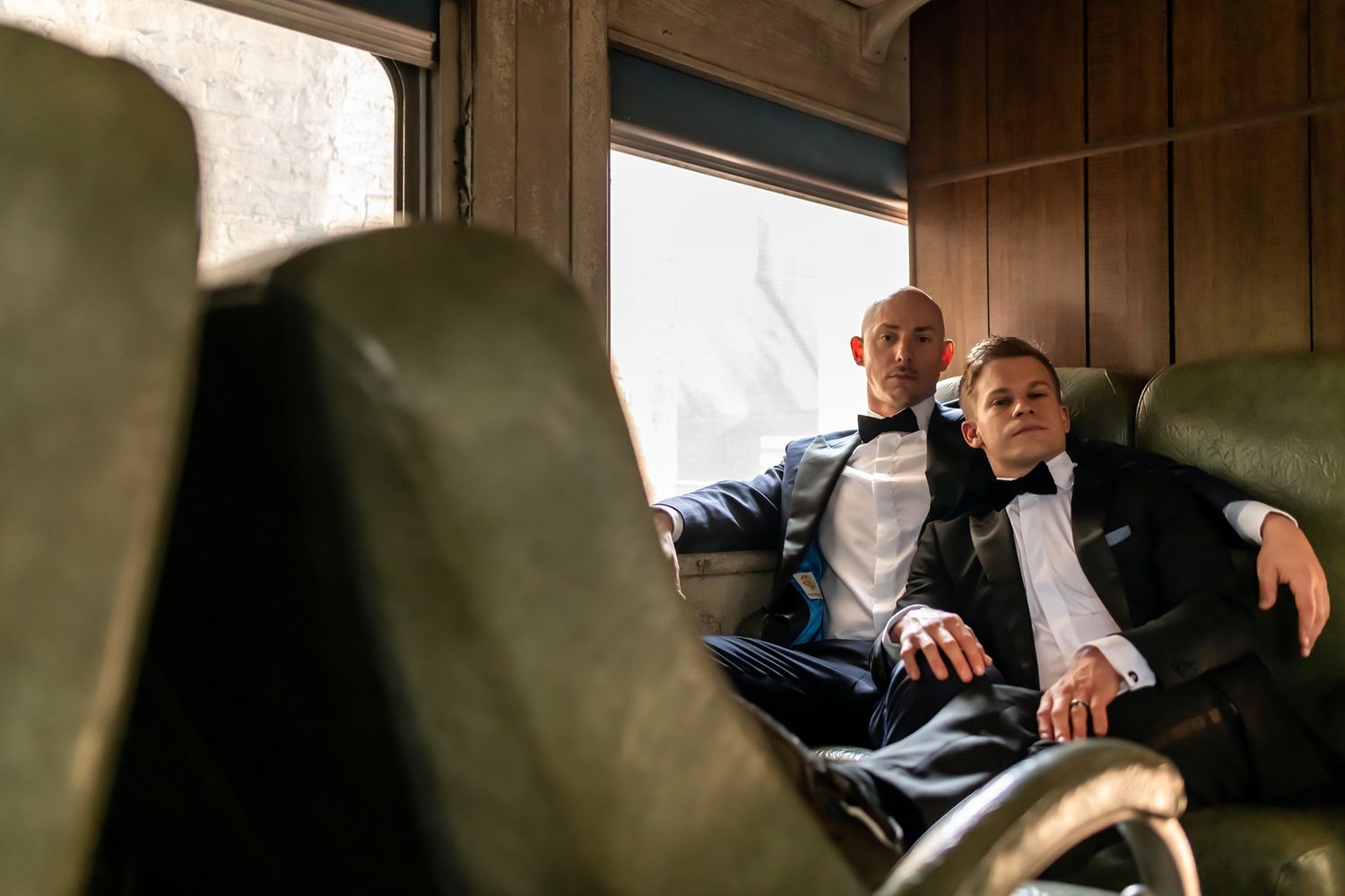 Two people in formal attire rest together on green leather seats inside what appears to be a luxury train car.