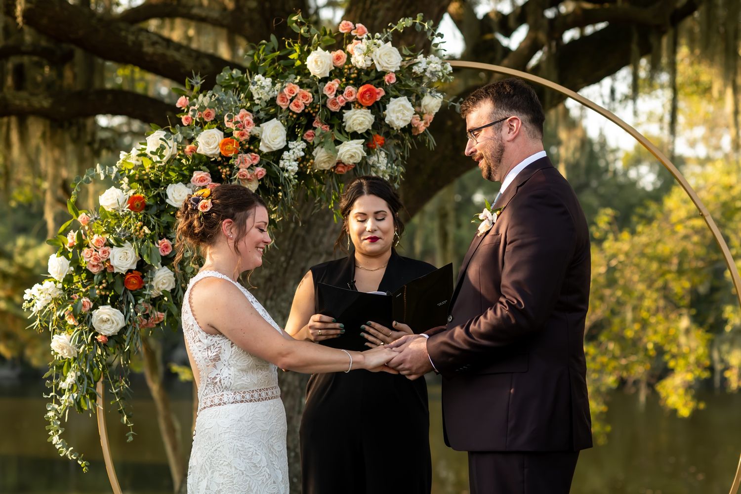 Couple exchanges vows under floral arch decorated with orange and white flowers during outdoor garden wedding ceremony.