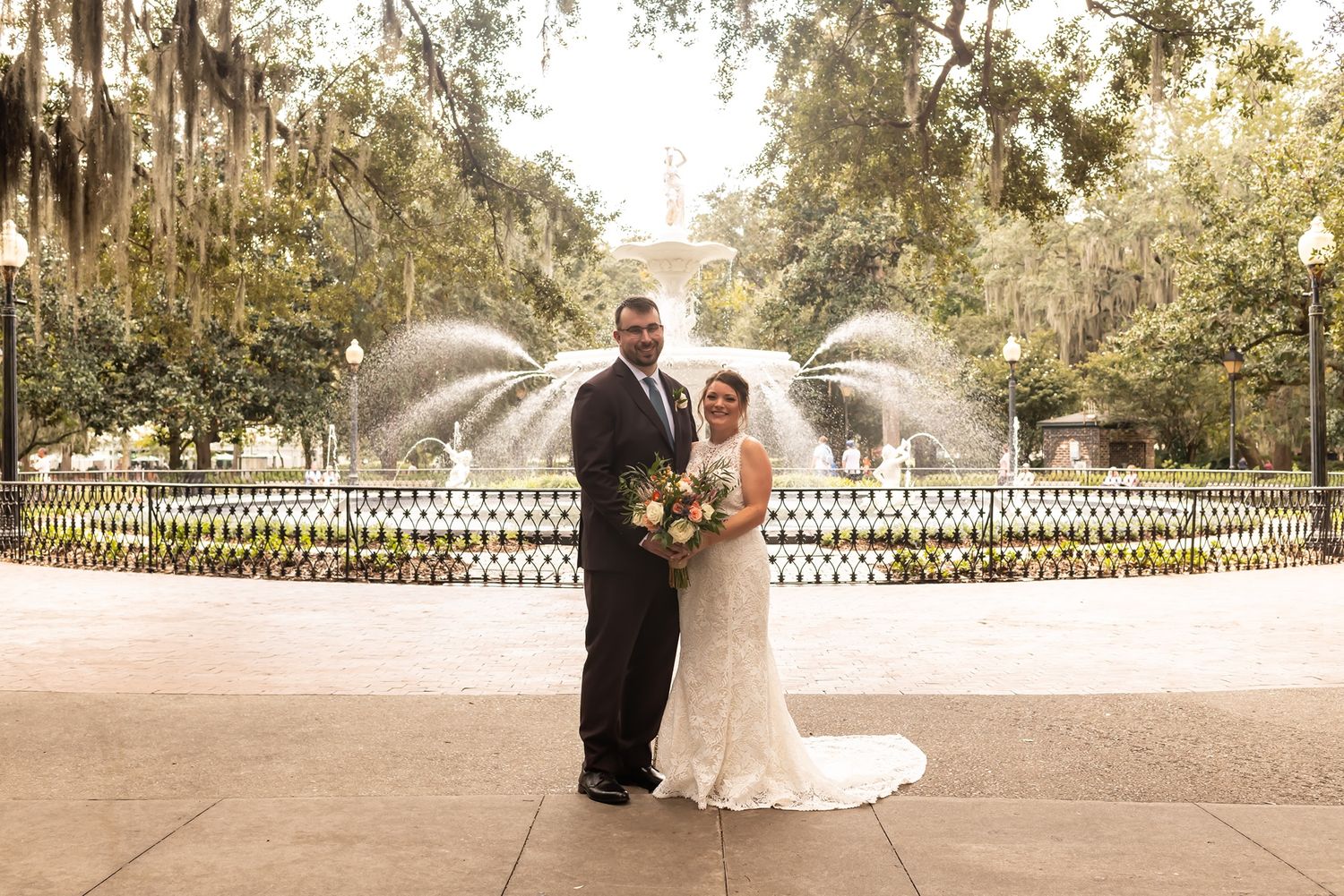 A wedding couple walking along a rustic path through wooded grounds with sunlight filtering through the trees.