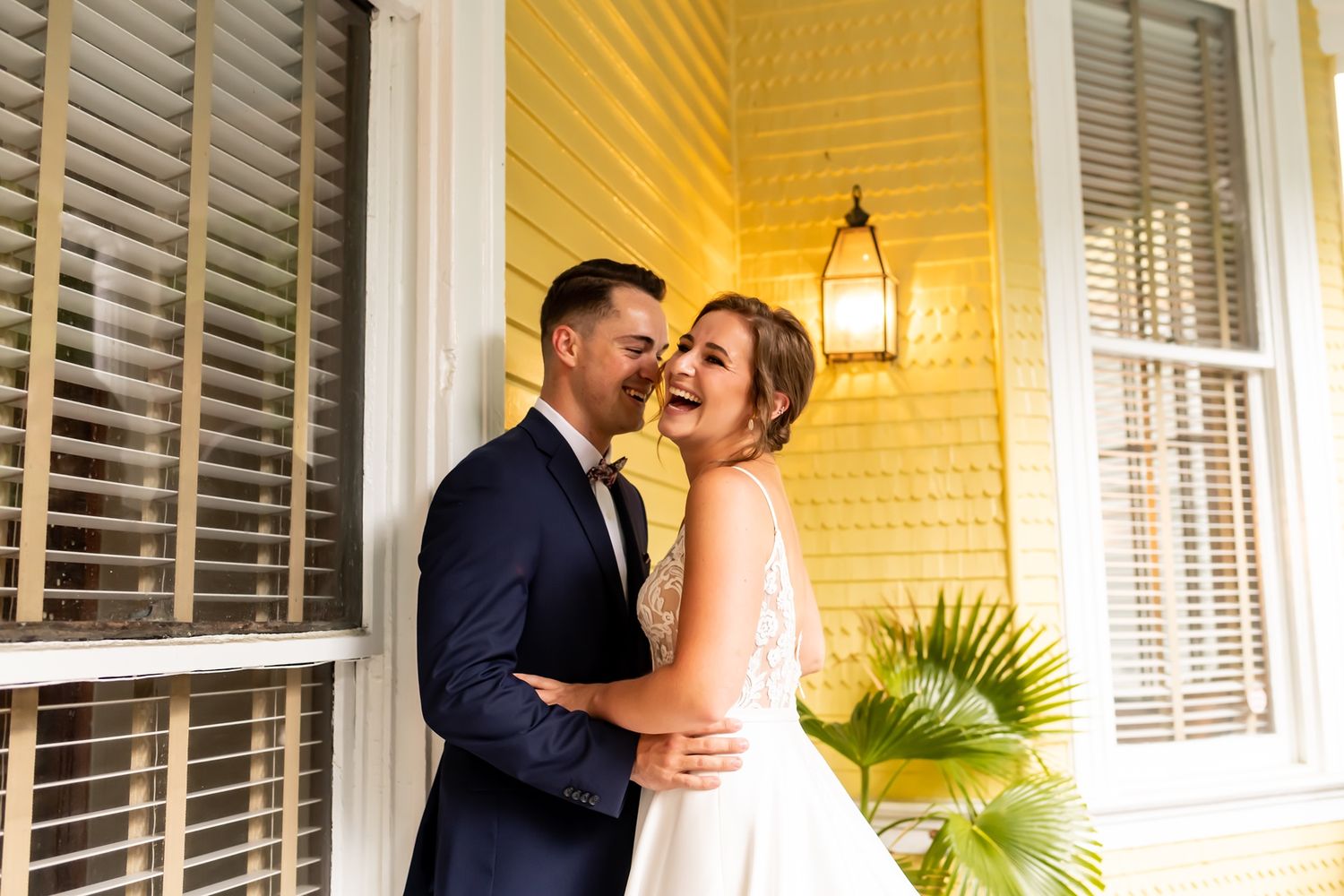 A couple embraces on a yellow porch with white shuttered windows and an outdoor lantern.