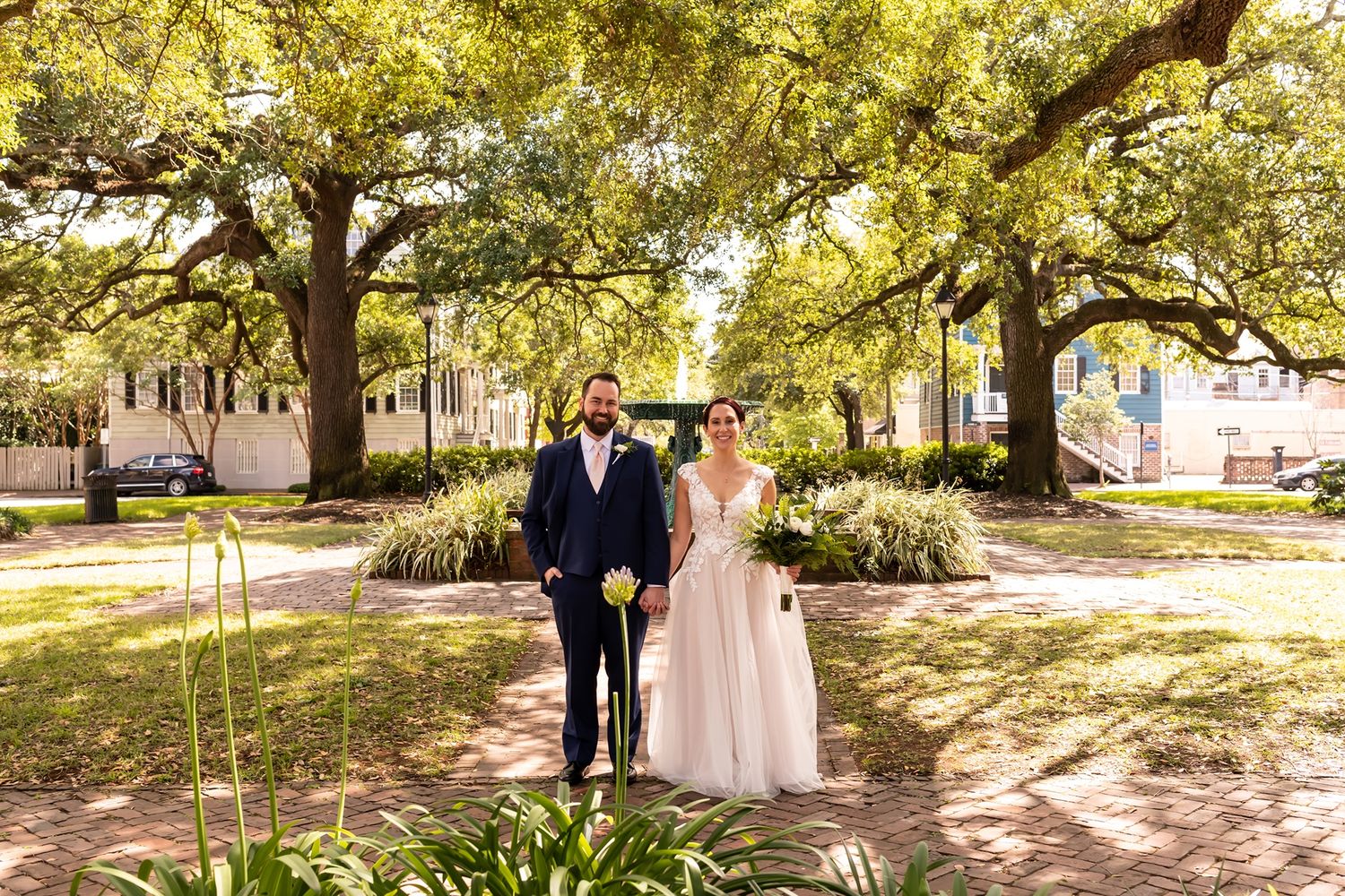 Couple in formal attire strolls along a scenic tree-lined pathway under golden sunlight.