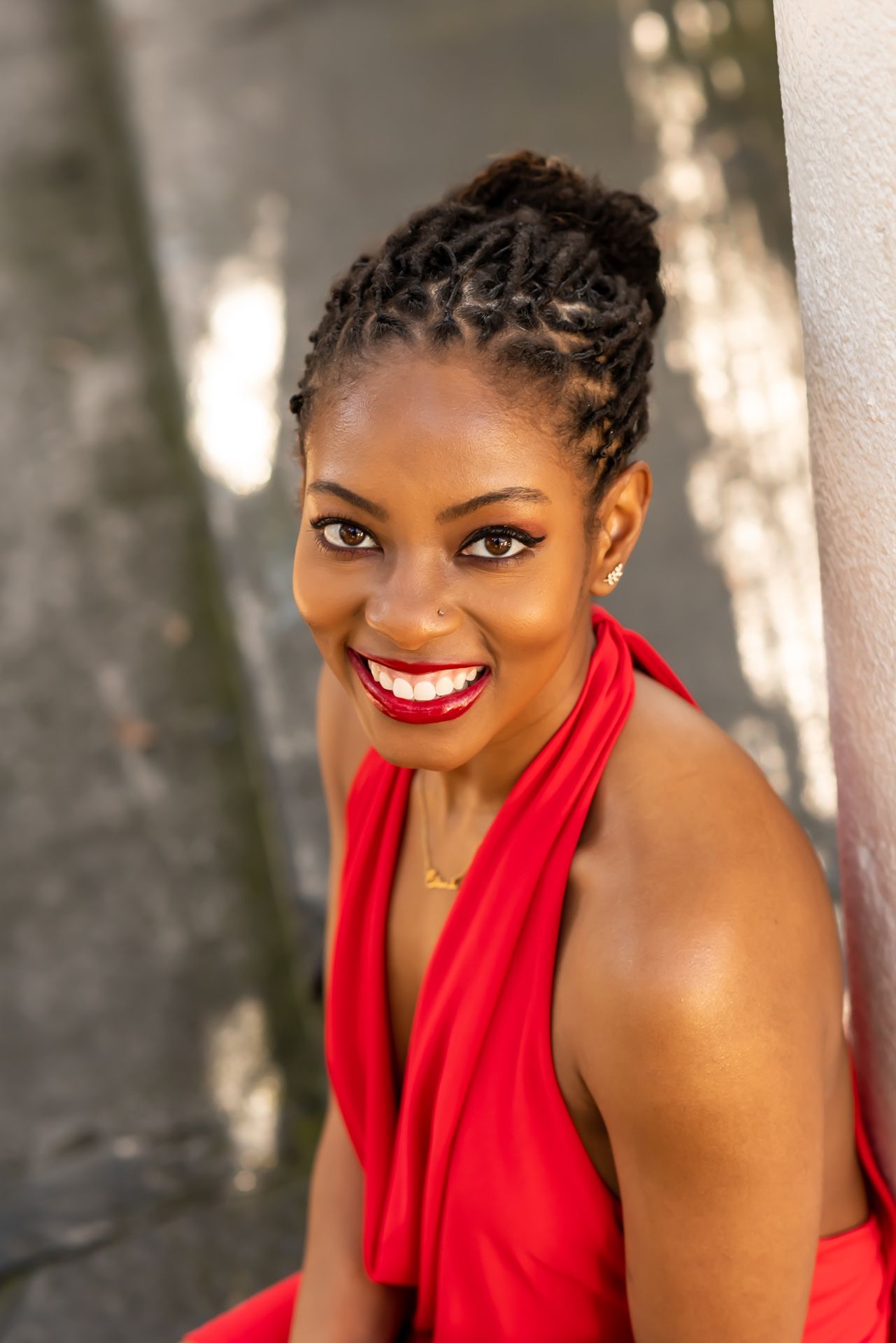 A figure in a vibrant red dress poses near a white column on a bright sunny day.
