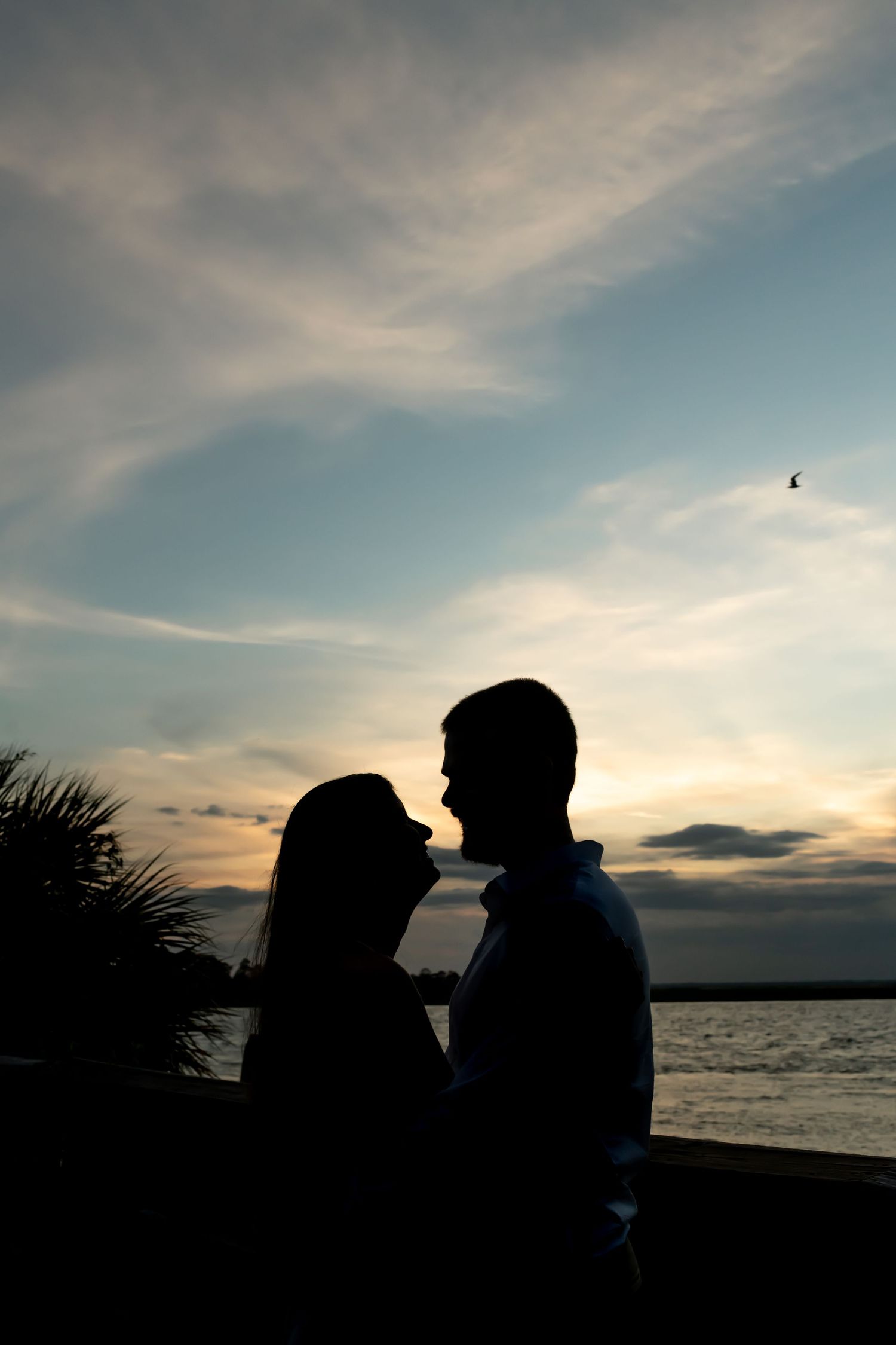 A silhouetted couple stands close together against a sunset sky over the ocean with palm trees and clouds in the background.